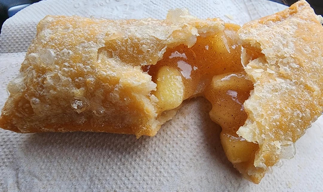 A close-up of a fried apple pie split open, revealing warm apple filling inside. The crispy, golden-brown crust sits on a textured, white paper napkin—a classic favorite among fast-food desserts.