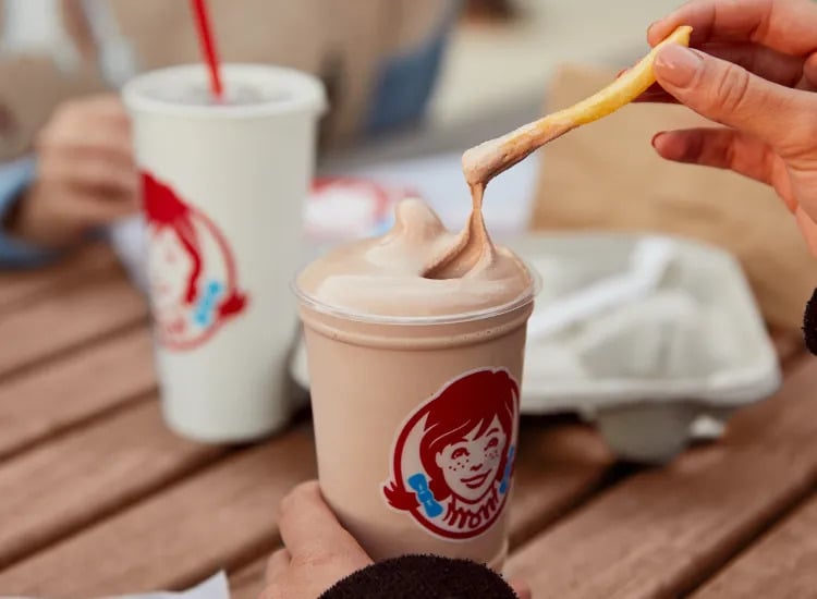 A hand dips a French fry into a chocolate Frosty—one of Wendy’s classic fast-food desserts—as another person holds a drink cup with a red straw at an outdoor table.