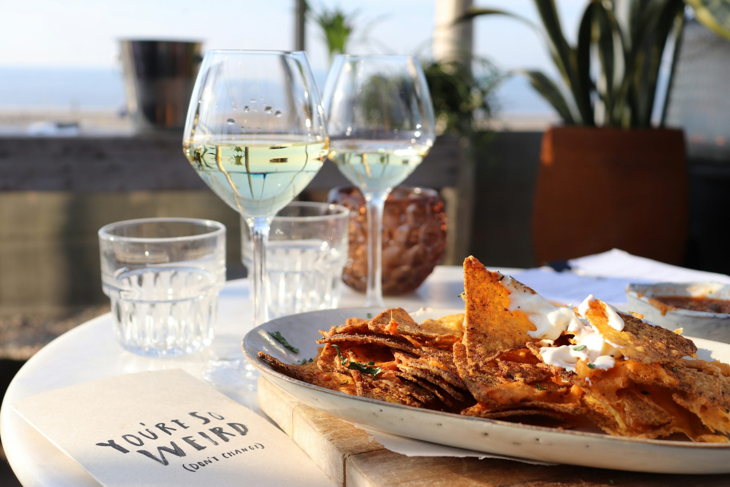A plate of nachos topped with sour cream sits on a table beside two glasses of white wine, two water glasses, a plant, and a napkin that reads "You're So Weird (but I like it)" with an outdoor, beachside view in the background.