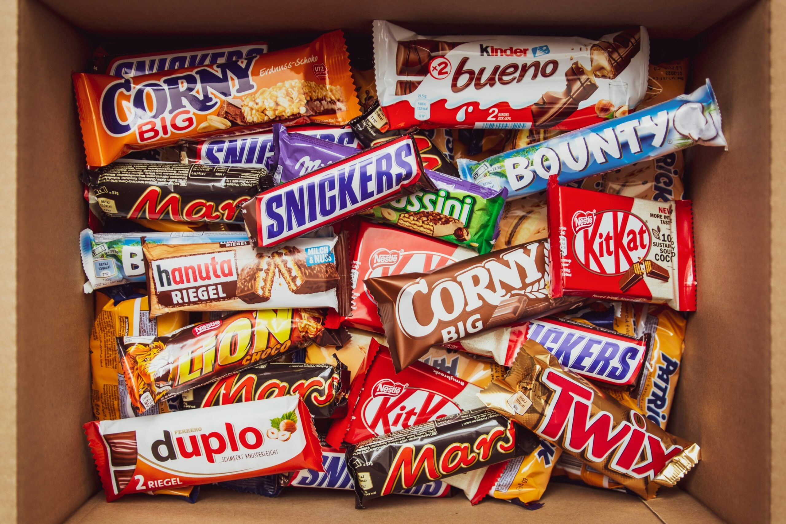 An open cardboard box filled with various assorted chocolate bars and candy bars, including brands like Snickers, Kinder Bueno, Bounty, KitKat, Mars, Twix, and Corny, arranged in a colorful, overlapping pile.