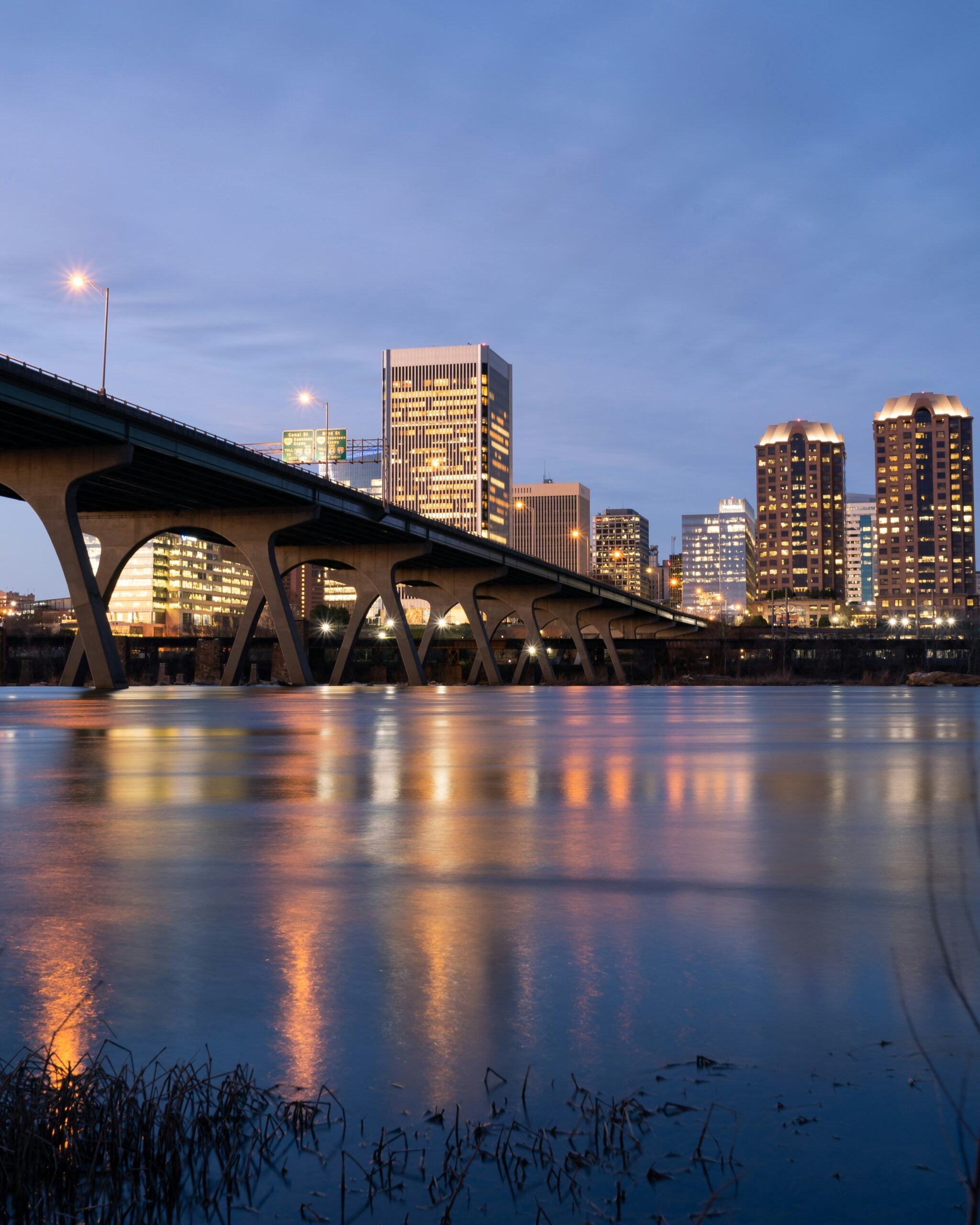 A city skyline at dusk with illuminated buildings and a large bridge spanning over a calm river, reflecting the city lights on the water’s surface.