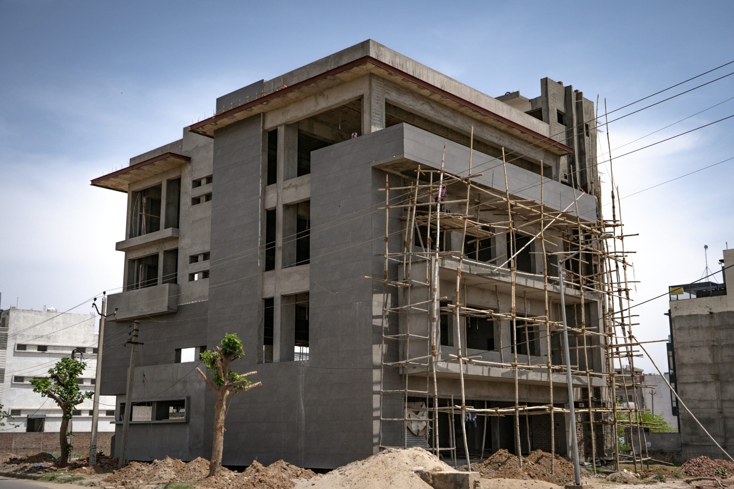 A multistory concrete building under construction with bamboo scaffolding along one side, reflecting ongoing urban development driven by Cost of Living Relief. Dirt piles, small trees, and power lines are visible beneath a clear sky.