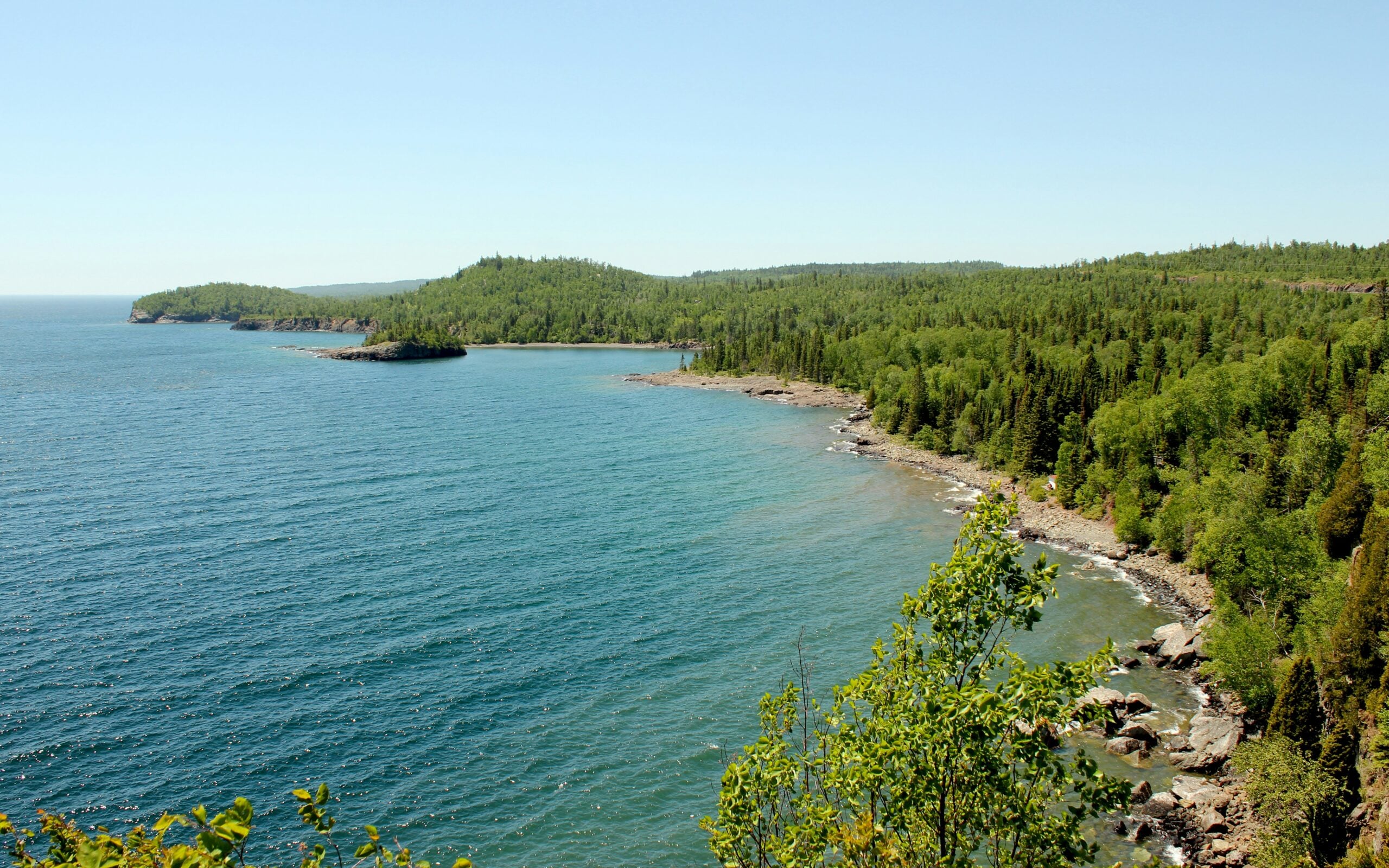 A scenic view of a large blue lake with a rocky shoreline and dense green forest, under a clear blue sky. The coastline curves gently into the distance, surrounded by lush trees and calm water.
