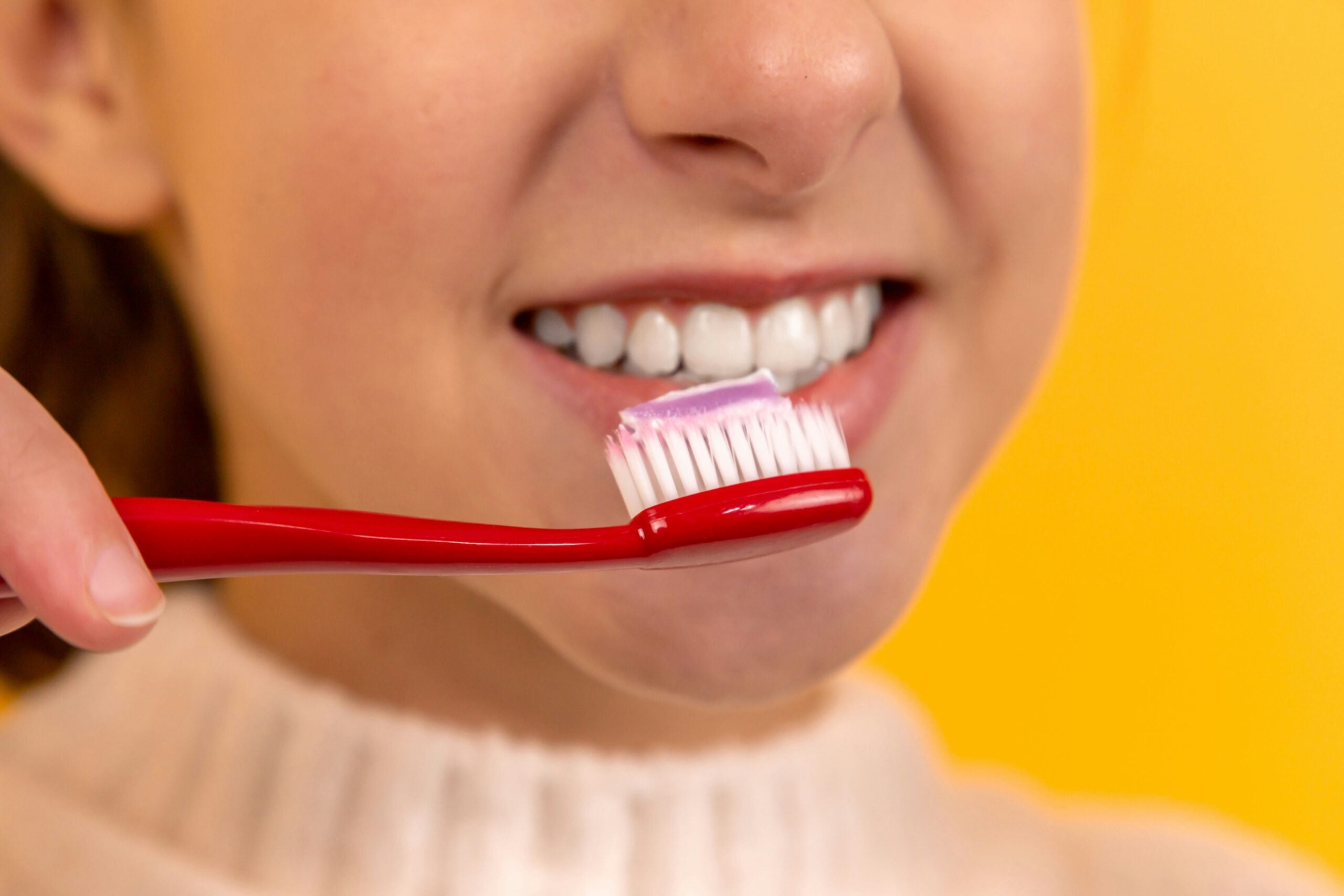 A person smiling and holding a red toothbrush with toothpaste on it near their teeth, against a bright yellow background.