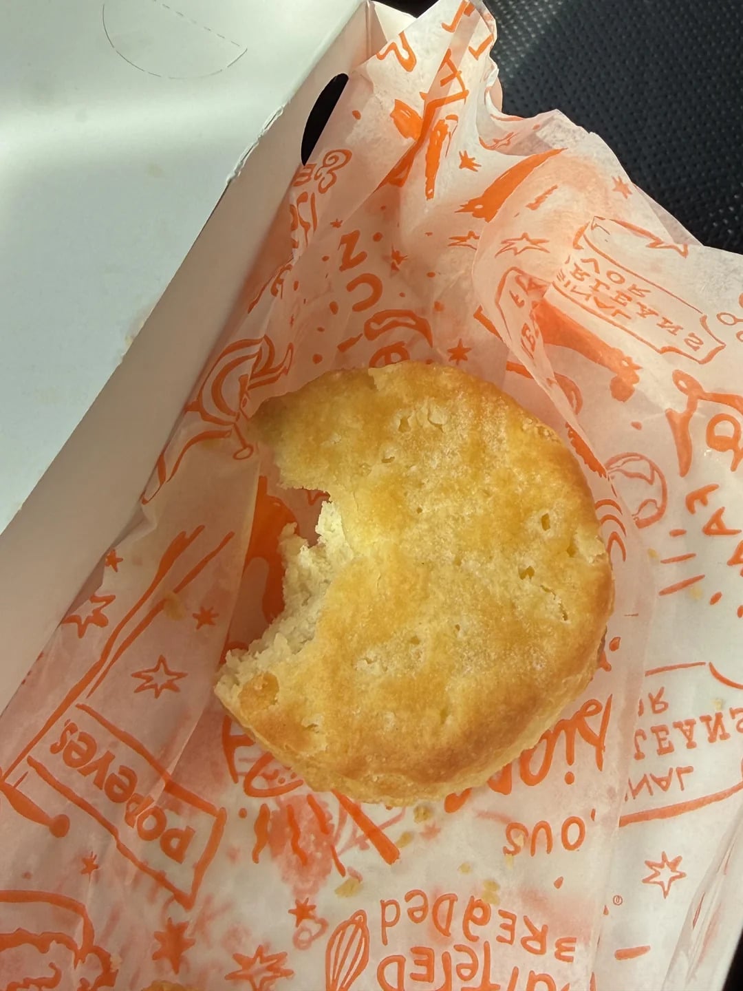 A golden-brown biscuit with a bite taken out sits on orange Popeyes-branded paper, next to a white takeout box.