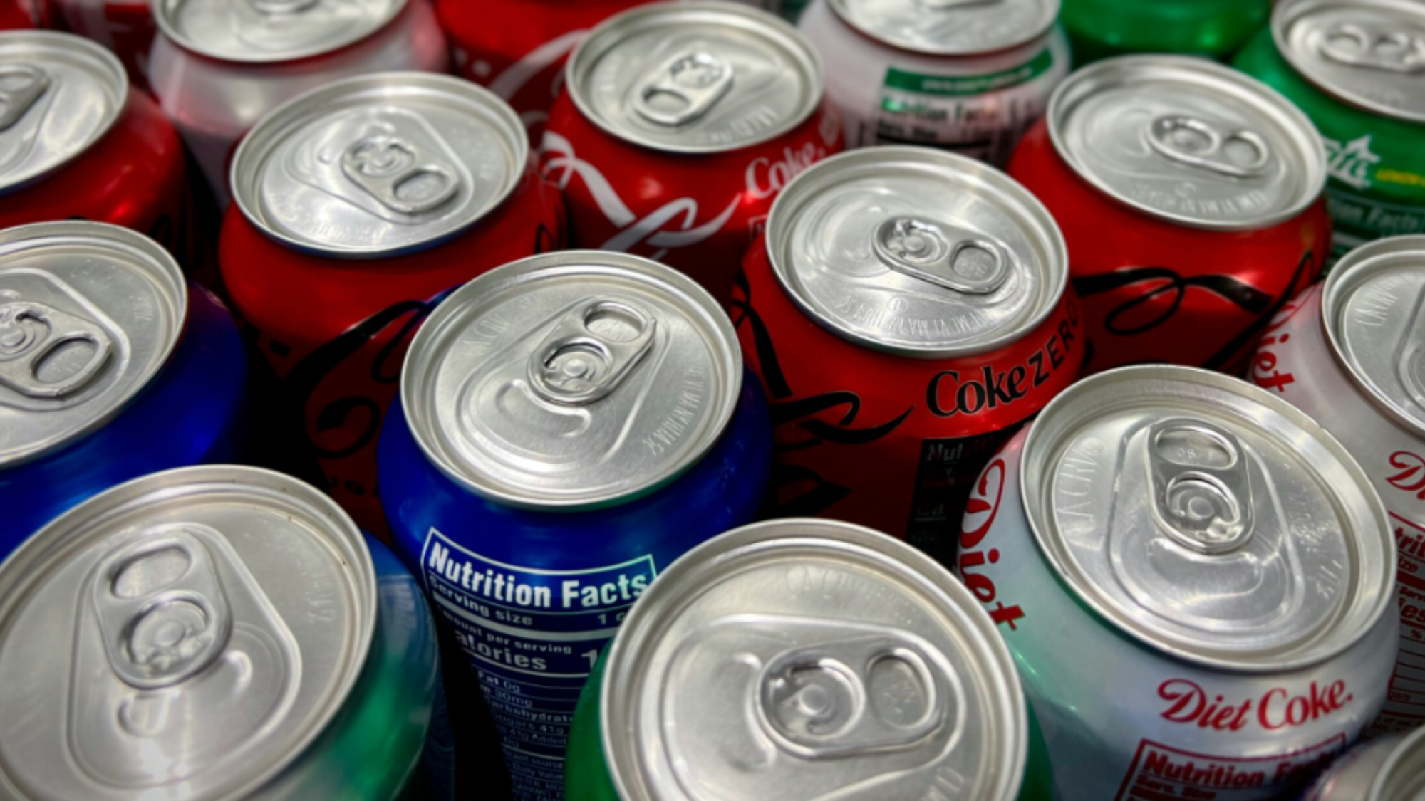 A close-up view of several unopened soda cans grouped together, featuring the best diet soda brands and flavors in red, blue, green, and silver colors with visible nutrition labels and pull tabs. Foods to never buy at full price.