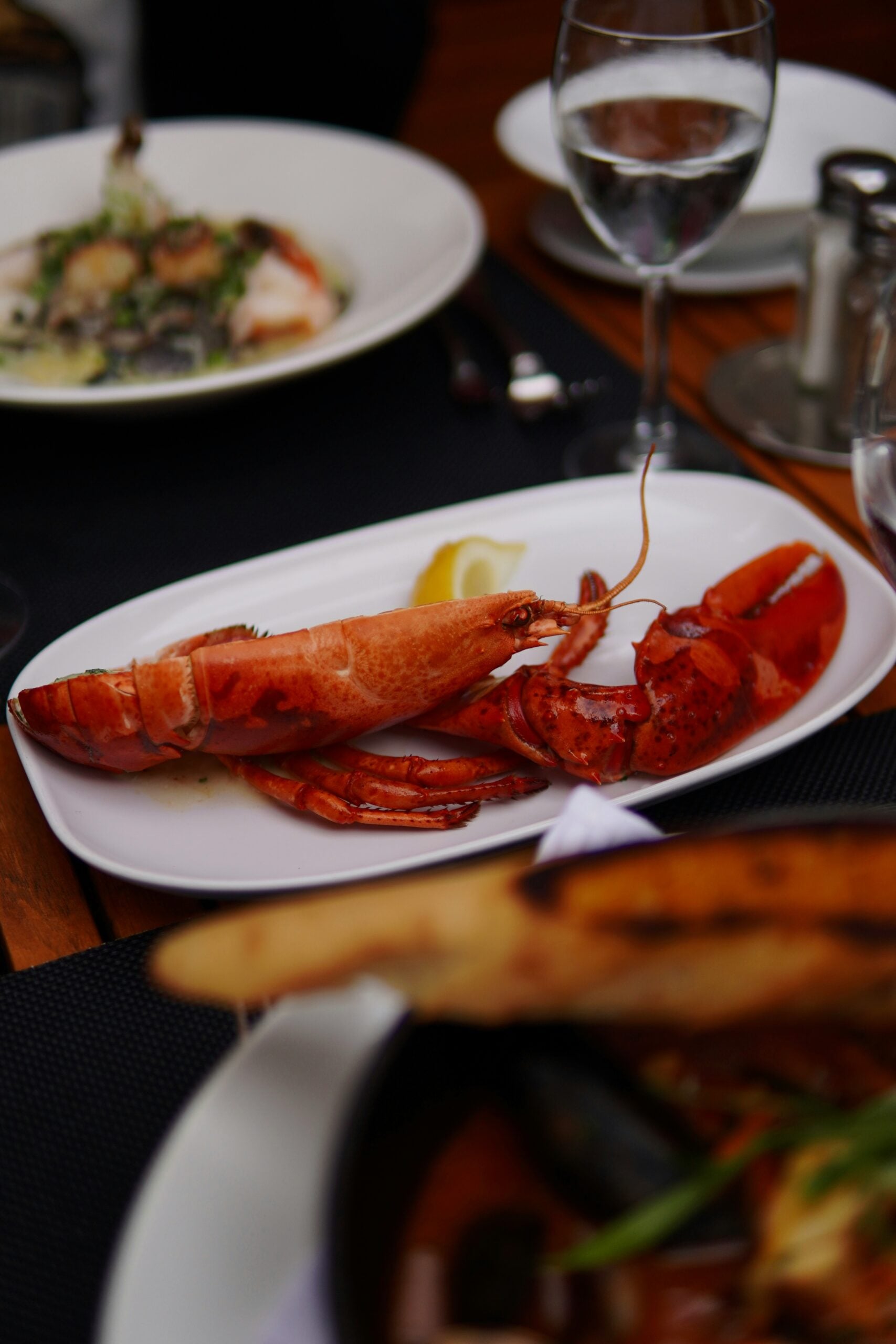 A cooked lobster is served on a white plate with a lemon wedge, accompanied by wine glasses and other dishes on a restaurant table.