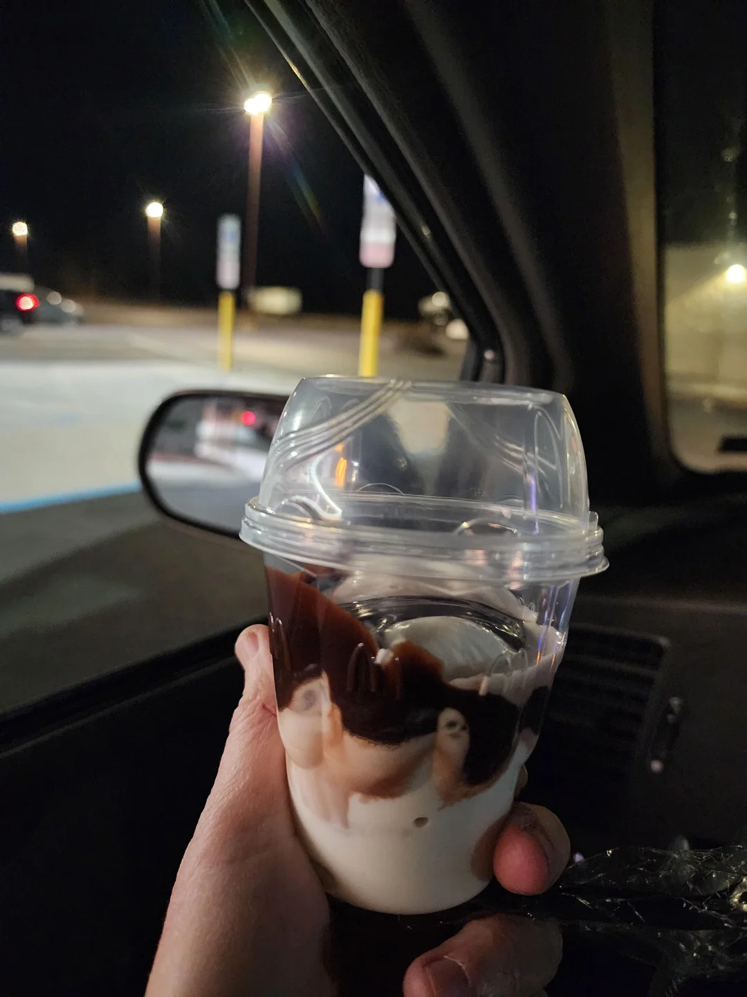 A hand holding a cup of vanilla soft serve ice cream topped with chocolate syrup inside a car at night, with streetlights and parking lot visible outside the window.