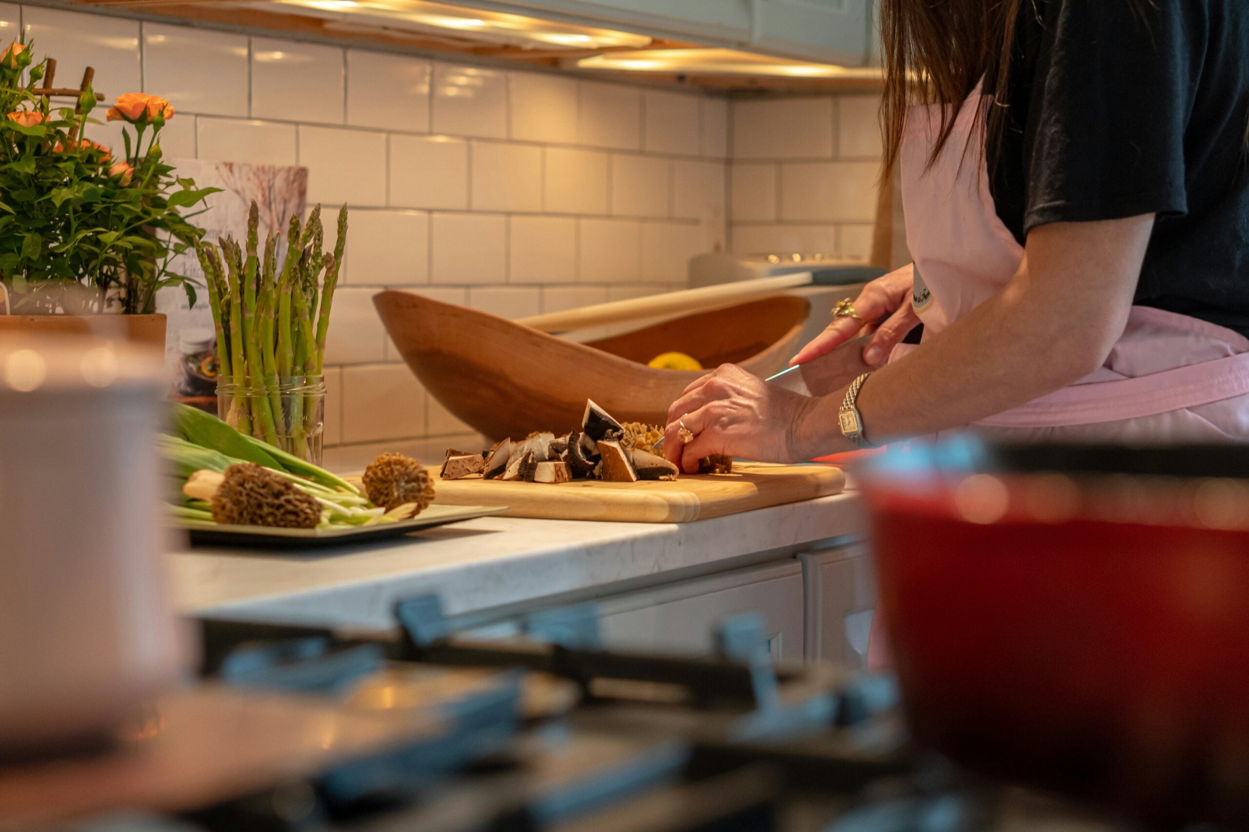 A person wearing a pink apron chops mushrooms on a wooden cutting board in a kitchen, with fresh vegetables and flowers visible on the counter nearby.