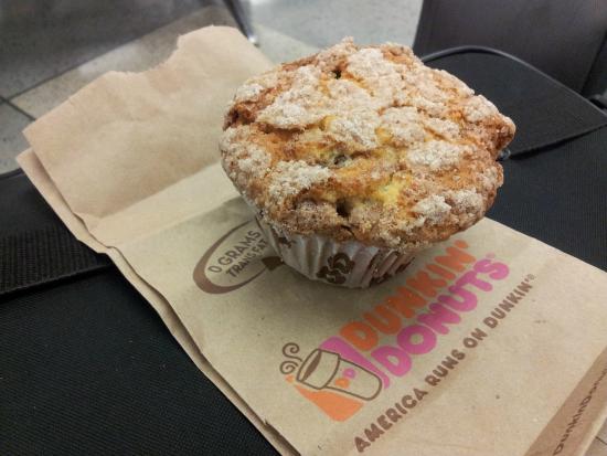 A crumb-topped muffin sits on a brown Dunkin' Donuts bag, which is placed on a black surface. The bag features the Dunkin' Donuts logo and slogan in orange and pink.