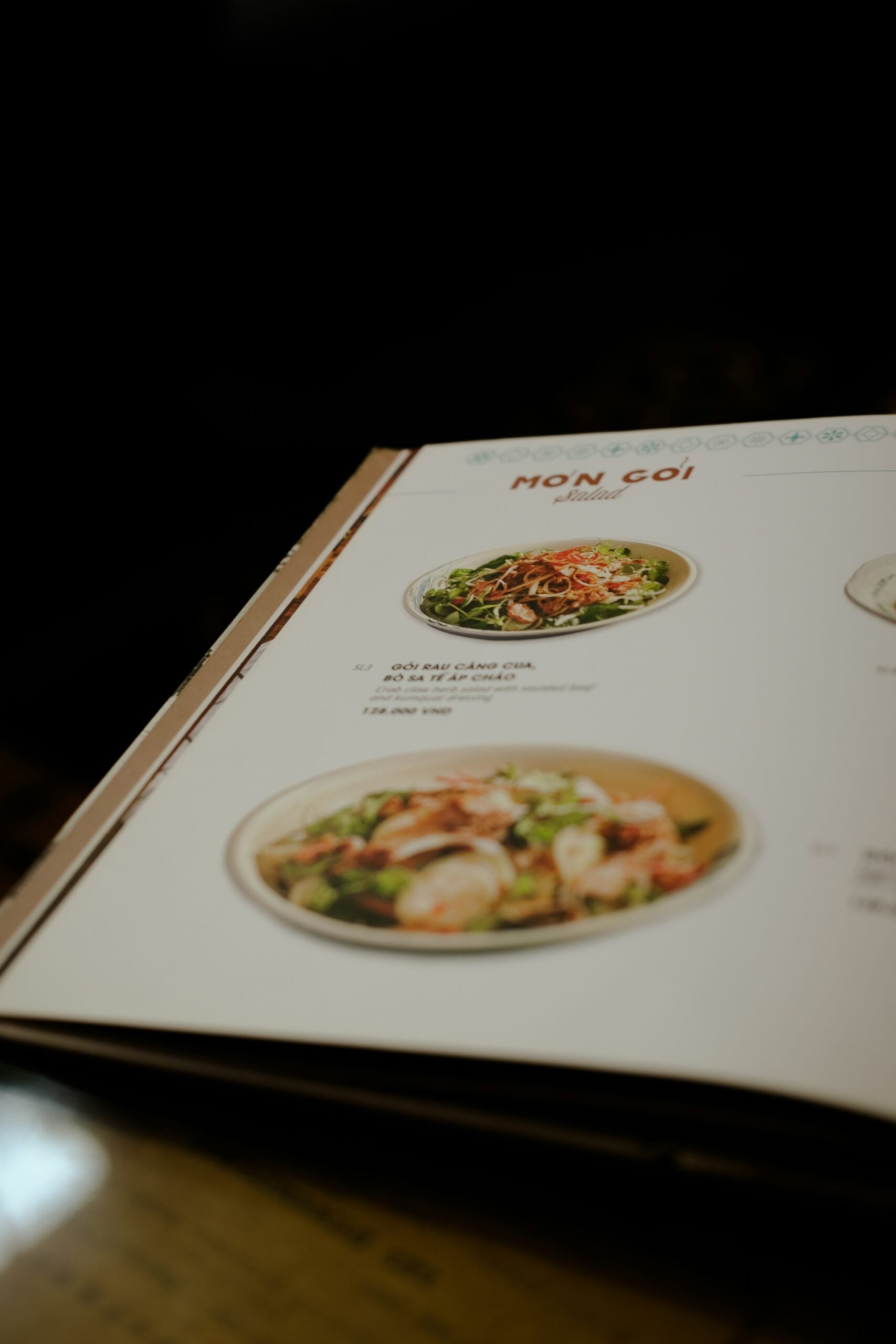 A close-up of a restaurant menu showing images and descriptions of two salad dishes, including ingredients and prices, with the heading "MON GOI" at the top. The menu is slightly open on a table.
