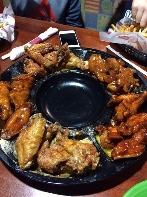 A platter of assorted chicken wings with various sauces and seasonings arranged around a black dipping bowl, with fries, a phone, and napkins visible on the table in the background.