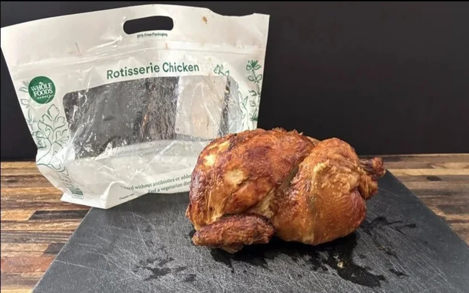 A delicious Rotisserie Chicken rests on a black cutting board in front of an empty Whole Foods rotisserie chicken bag, set against a wooden surface and black background.