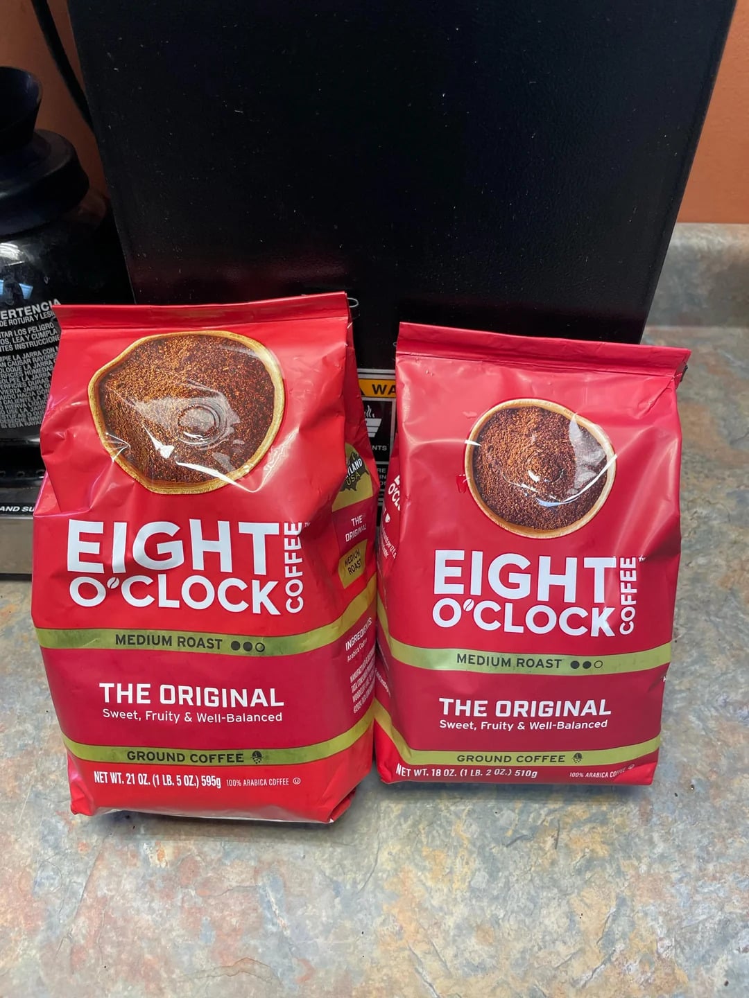 Two red bags of Eight O'Clock Coffee, one of the classic coffee brands, labeled "The Original," sit side by side on a kitchen counter in front of a black appliance. The Medium Roast bags are 21 oz and 18 oz in size.