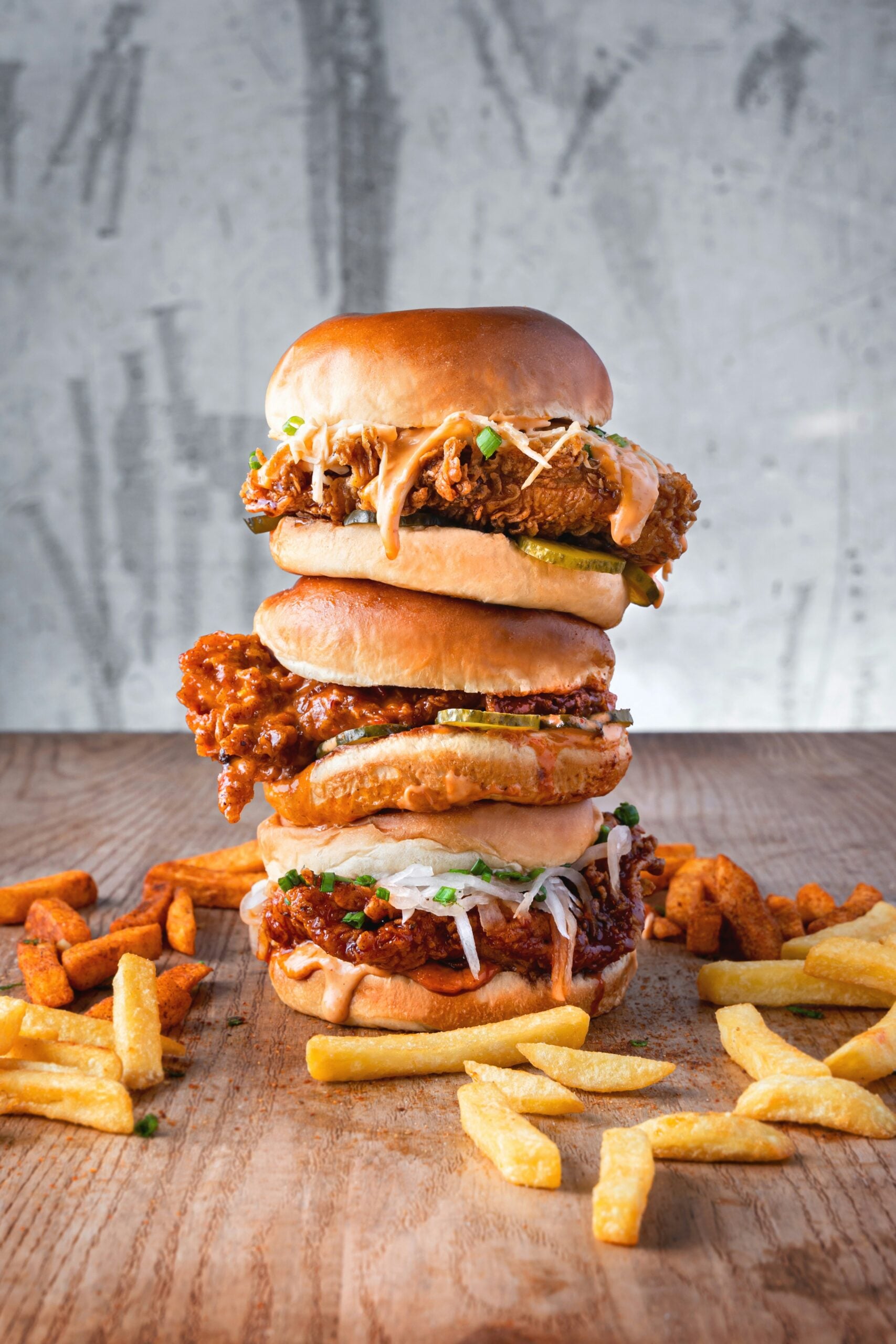 Three stacked fried chicken sandwiches with various toppings, surrounded by scattered French fries and waffle fries on a wooden table against a textured gray background.