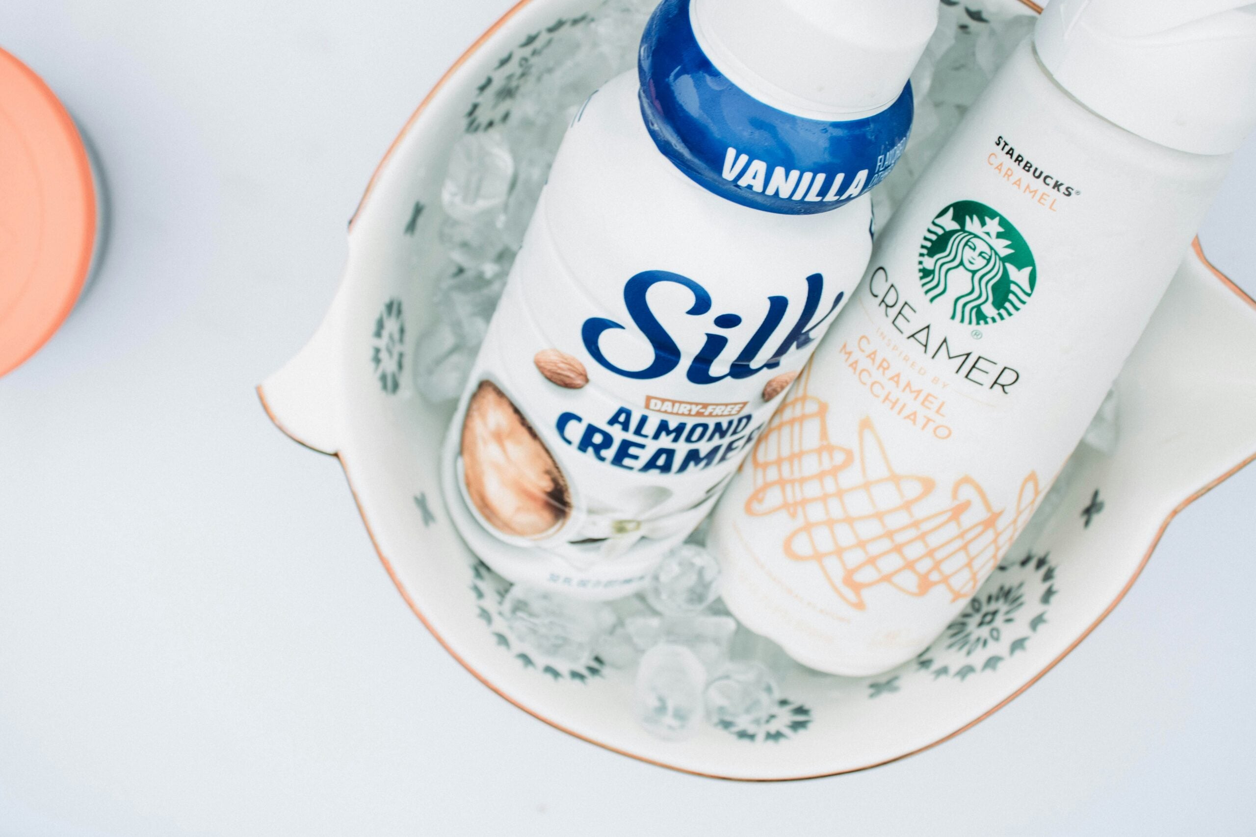 Two bottles of coffee creamer, Silk Almond Creamer (Vanilla) and Starbucks Caramel Creamer, sit in a bowl filled with ice cubes on a white surface.