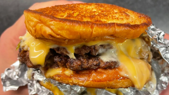 A close-up of a grilled cheese burger with melted cheese oozing over two beef patties, held in a hand wrapped with aluminum foil. This fast-food secret menu hack features a toasted golden brown bun on top and bottom.