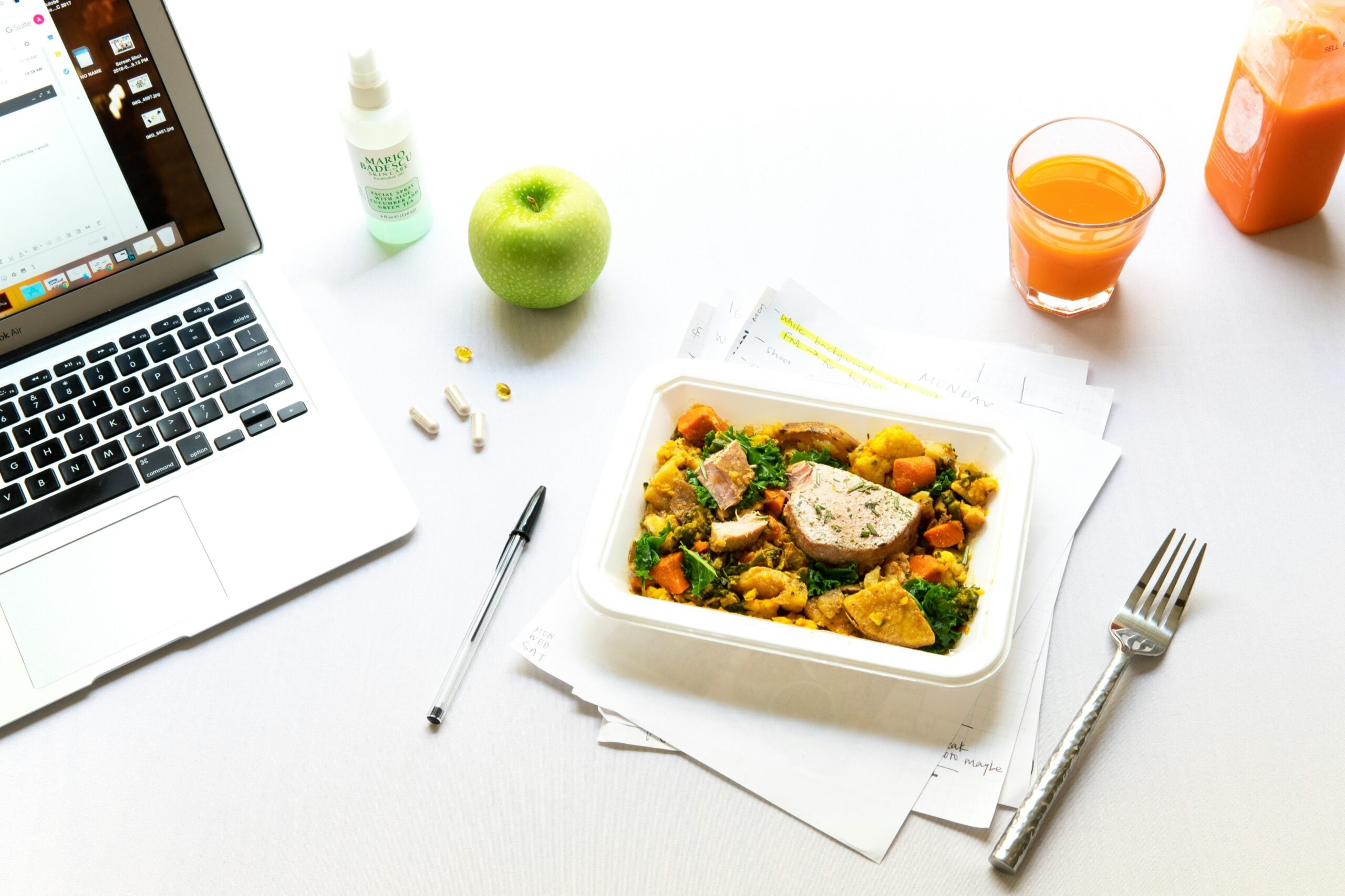 A workspace with a laptop, green apple, vitamins, pen, fork, orange juice, and a container of salad with grilled chicken on scattered papers, all on a white surface.