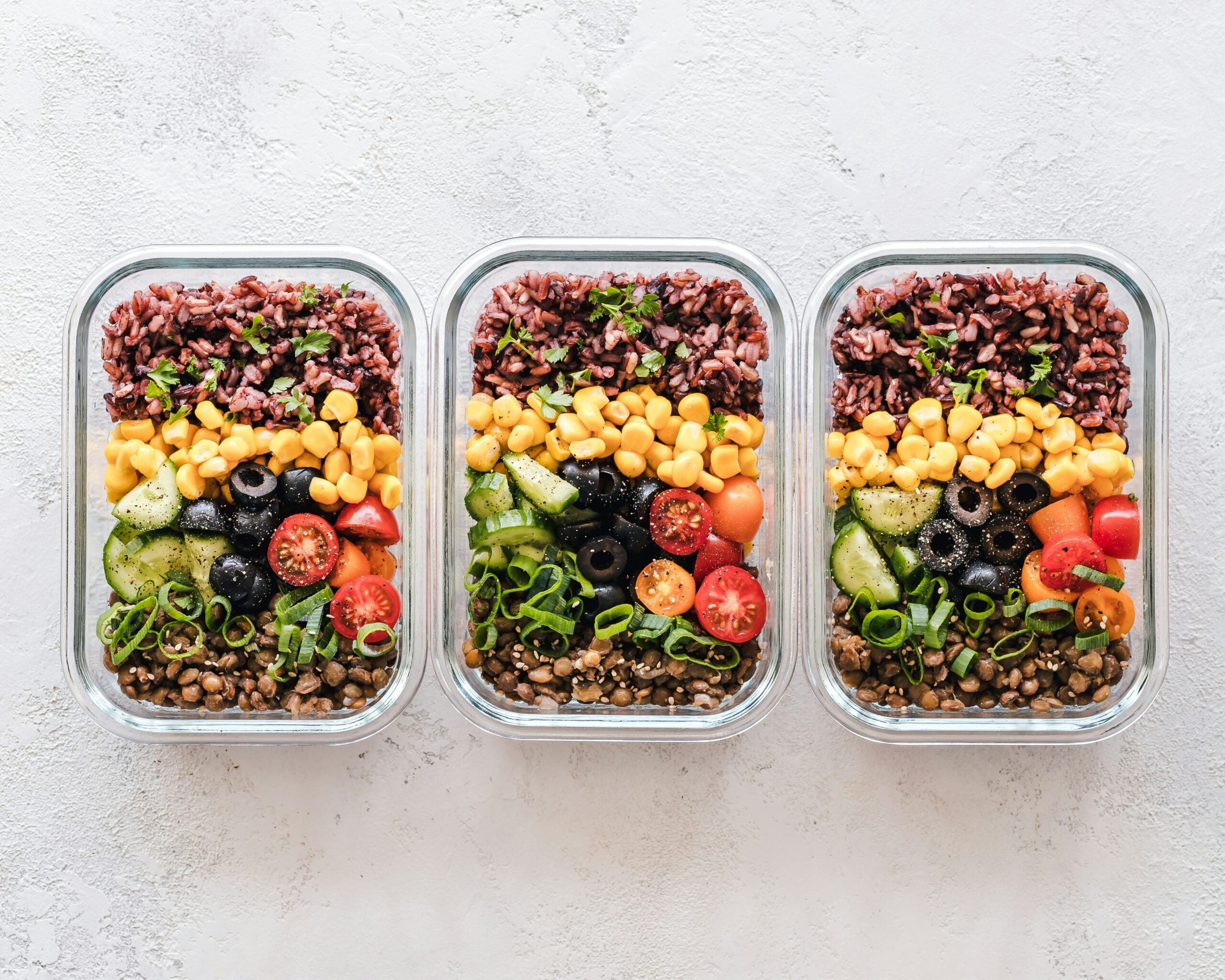 Three glass meal prep containers filled with colorful layered ingredients: brown rice, corn, cherry tomatoes, cucumber, black olives, green onions, and lentils, arranged neatly on a light textured surface.