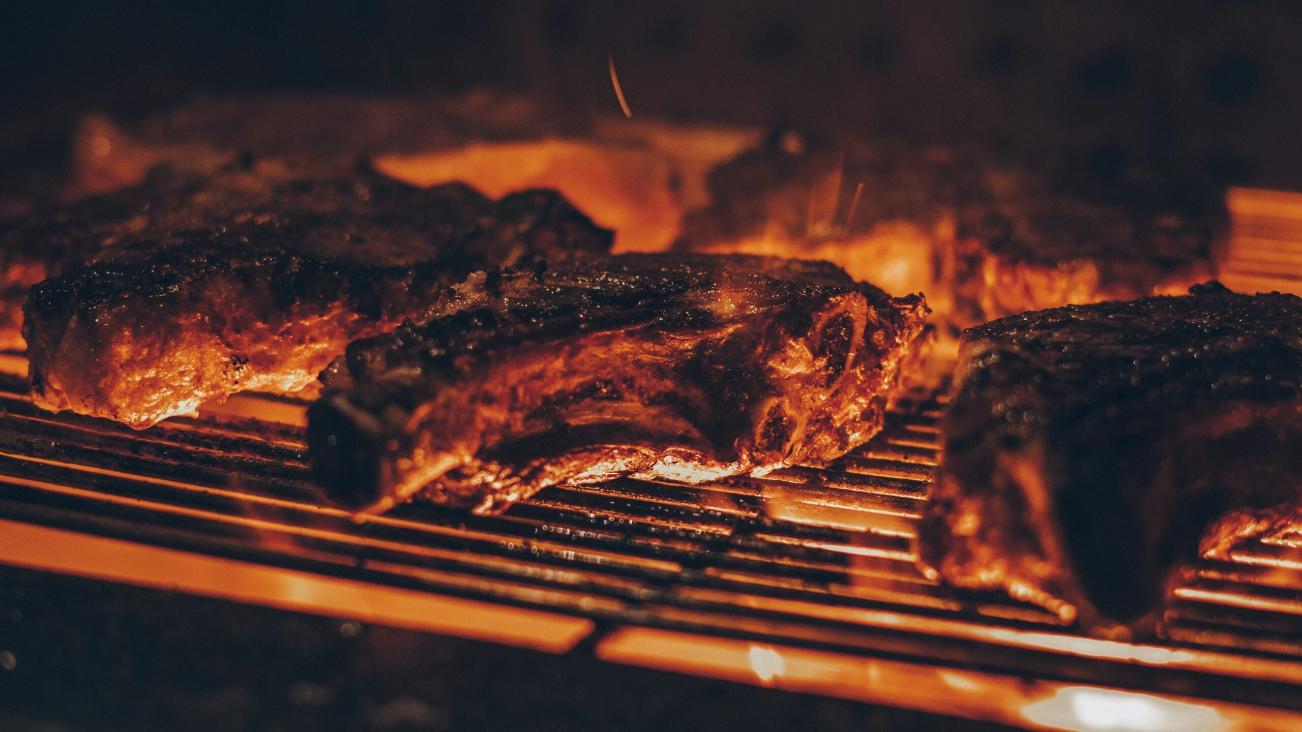 Thick, juicy steaks sizzling on a hot grill over open flames, with a warm, orange glow highlighting the charred edges and grill marks.