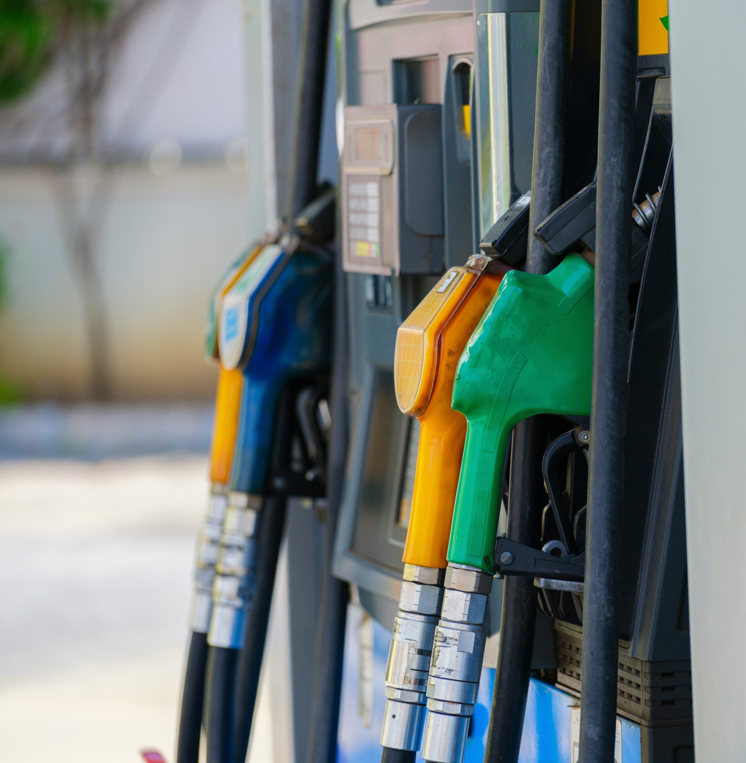 Close-up of fuel pump handles at a gas station, showing green, yellow, and blue nozzles attached to the dispenser, with a blurred background.