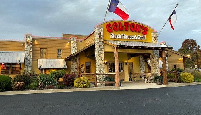 Exterior of Colton’s Steak House & Grill with a red and yellow sign, stone columns, wooden accents, two flags above the entrance, and landscaping in front on a cloudy day.
