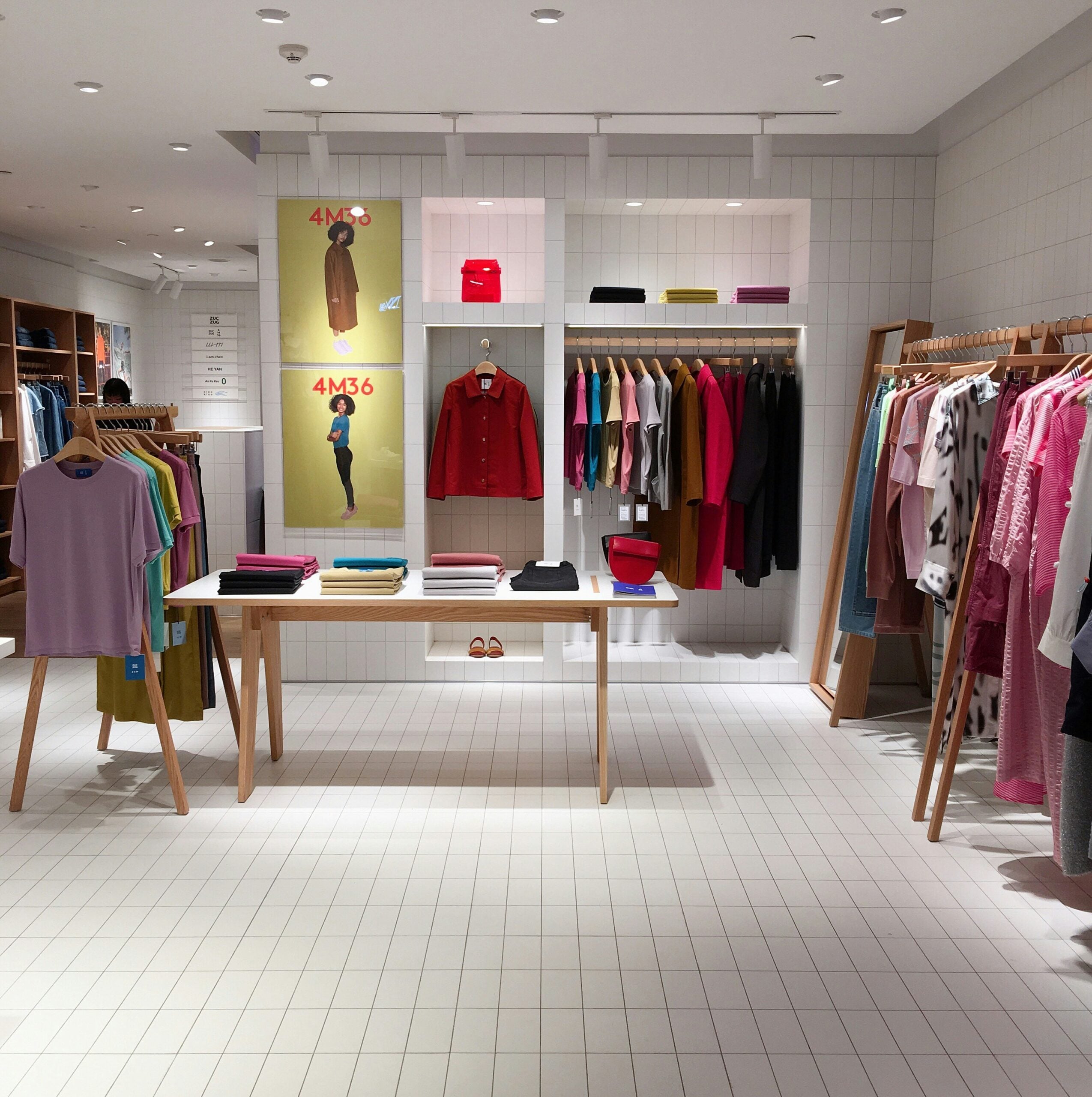 A modern clothing store with neatly arranged clothes on racks and shelves, a table displaying folded garments, and posters of models on the wall above a red shirt. The space is well-lit with a minimalist design.