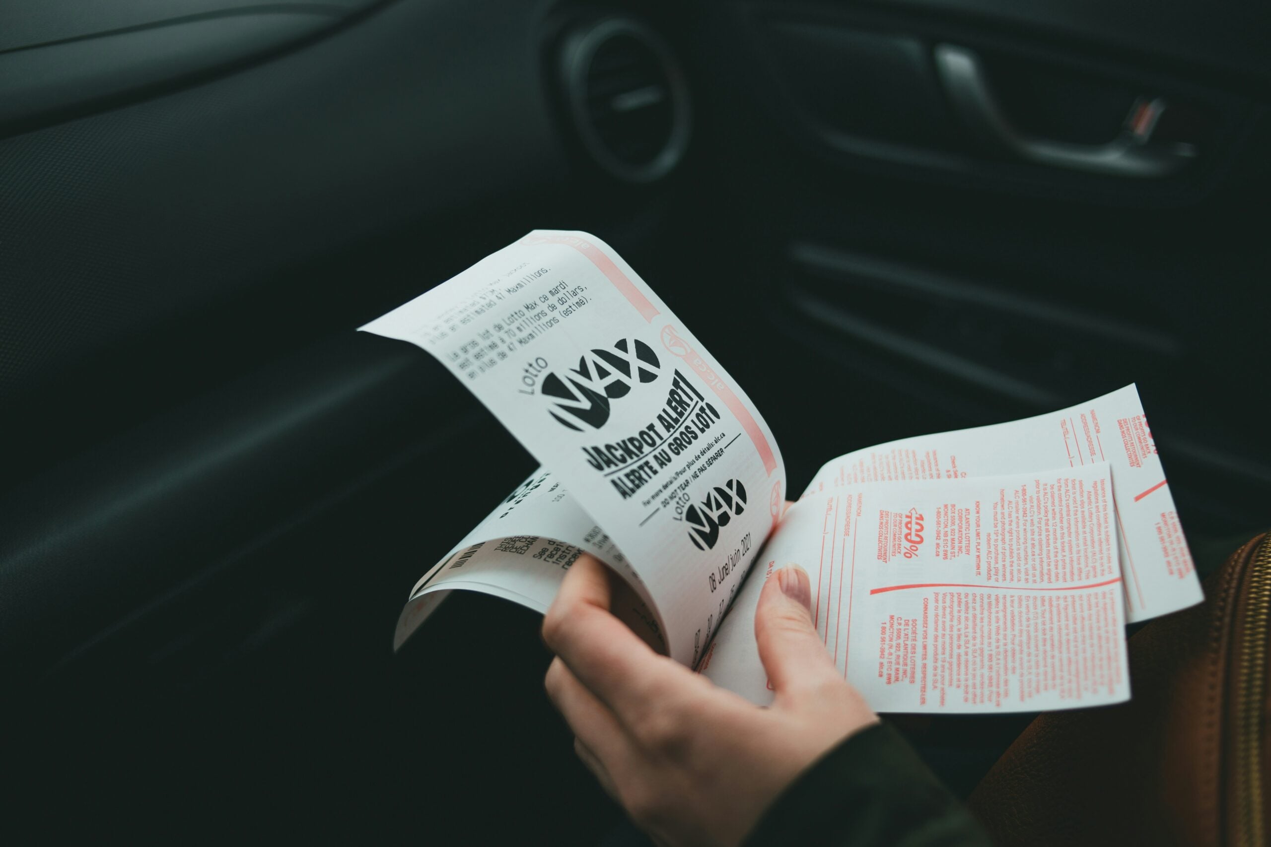 A person sitting in a car holds and looks at a folded pamphlet or brochure with bold black and red text and graphics on white paper. The car’s door and dashboard are visible in the background.