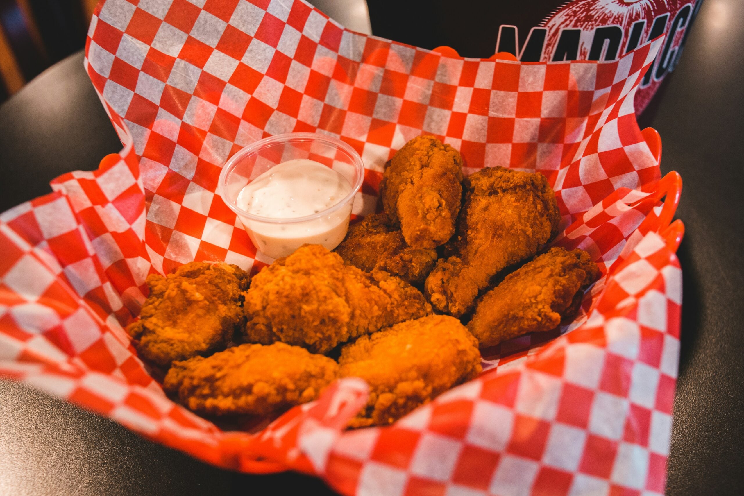 A basket lined with red and white checkered paper holds several pieces of fried chicken wings and a small container of ranch dipping sauce.