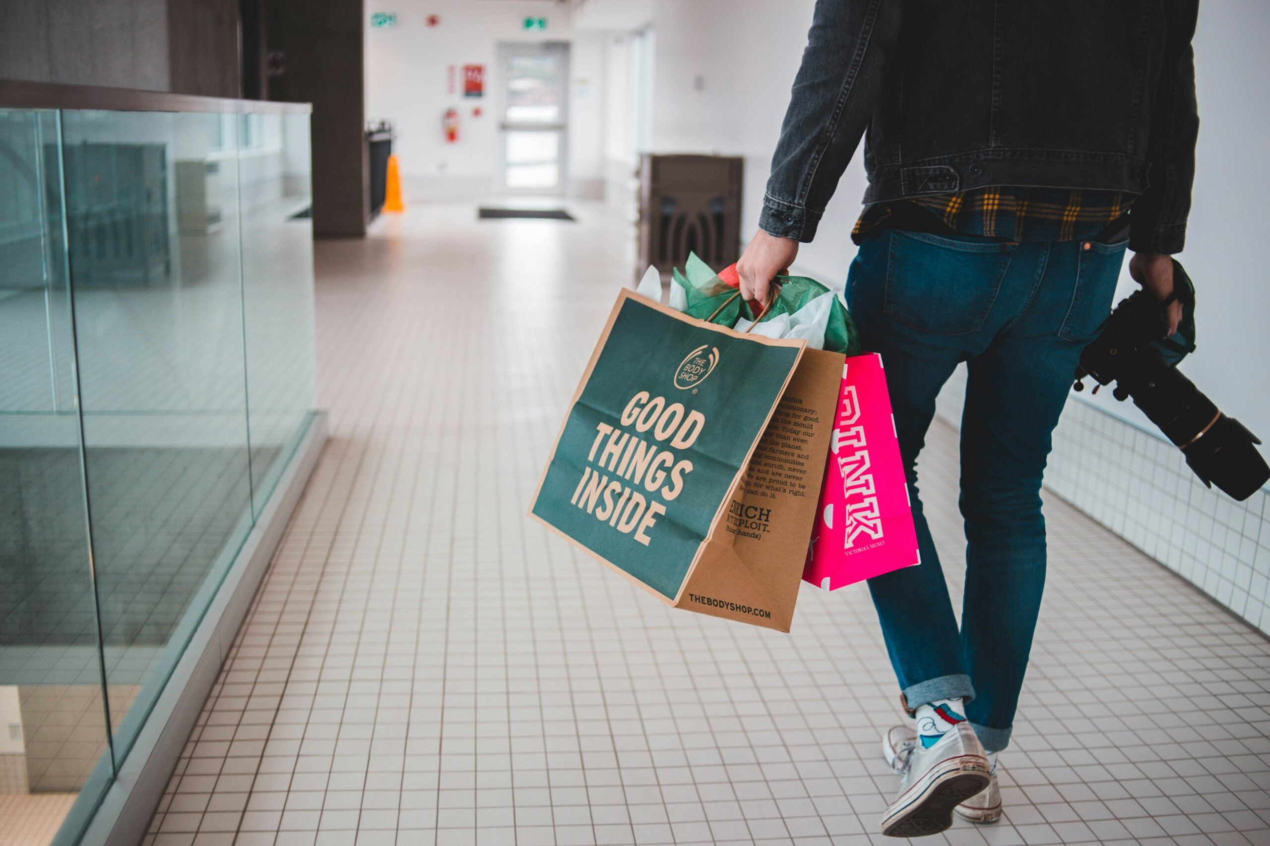 A person in jeans and a denim jacket walks down a tiled hallway, holding shopping bags and a camera in one hand. The bag in front reads "Good Things Inside.