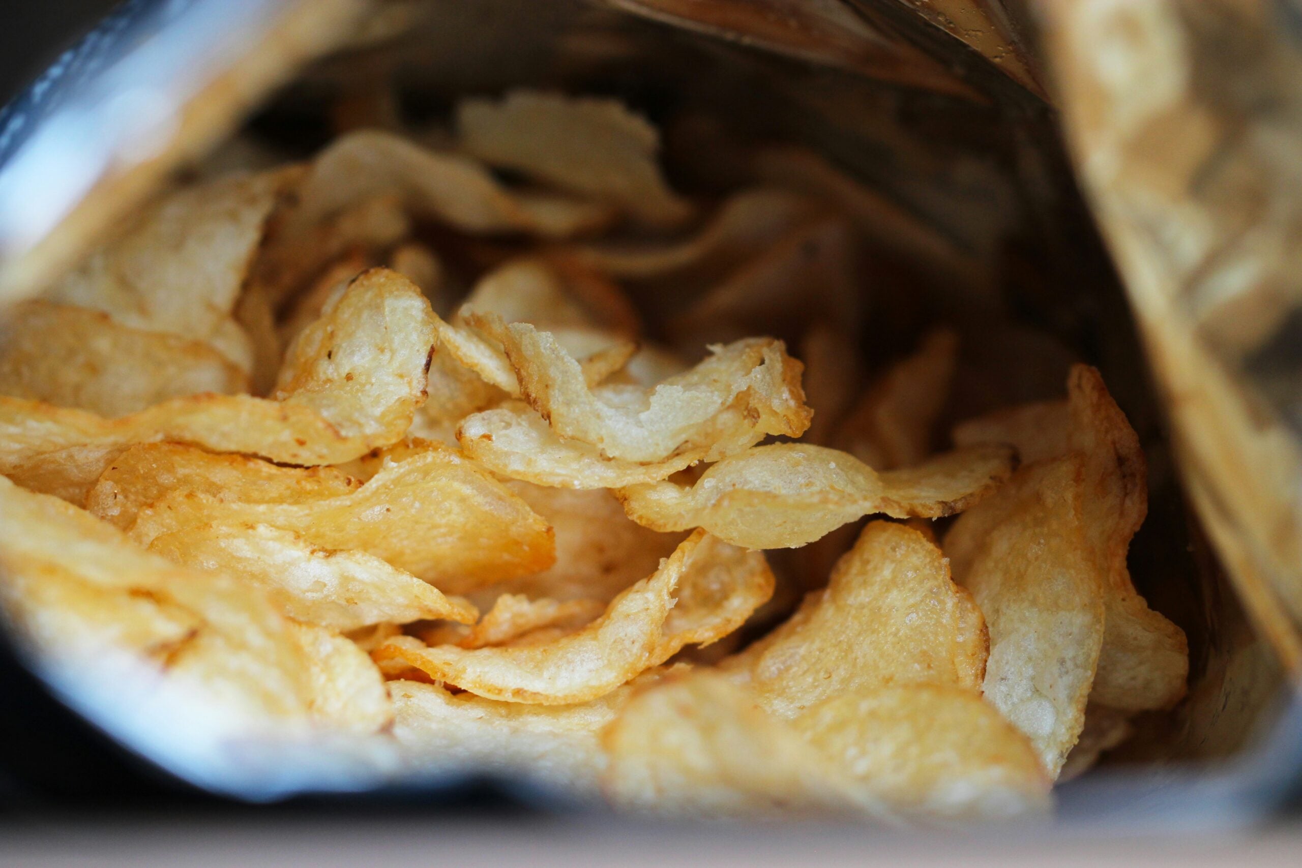 An open bag of potato chips showing a close-up view of the crispy, golden chips inside. The chips have a wavy texture and are surrounded by the reflective interior of the bag.