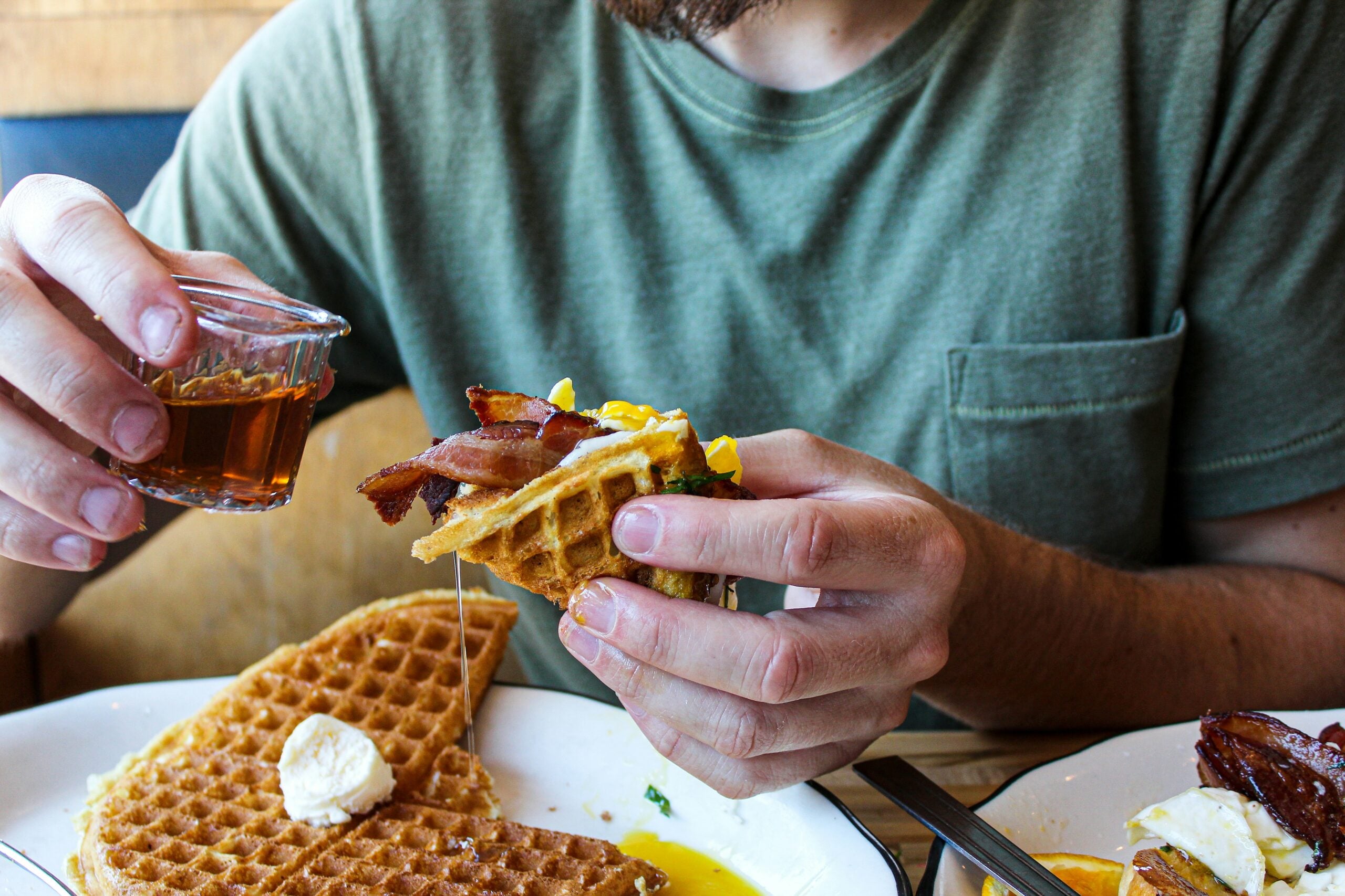 A person in a green shirt pours syrup onto a waffle topped with bacon and eggs, holding it in one hand over a plate with more waffles and butter.