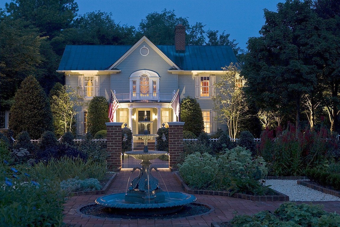 A two-story, light blue house with warm lights shines at dusk, surrounded by a lush garden. Reminiscent of Small-Town Luxury Hotels, American flags flank the entrance and a decorative fountain sits on a brick walkway in the foreground.