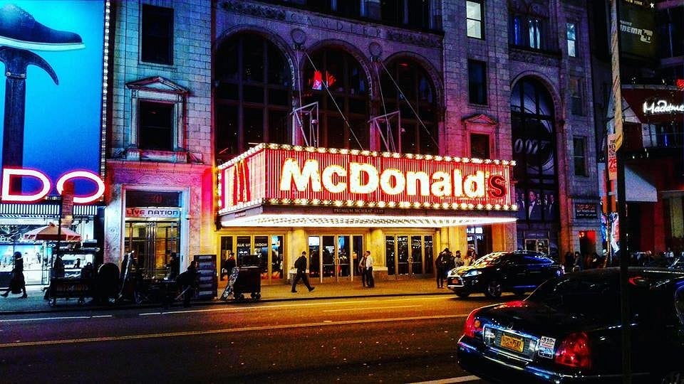A brightly lit McDonald’s restaurant with a large, illuminated marquee sign on a busy city street at night—one of the Coolest McDonald's Locations—surrounded by bustling people, cars, and neighboring buildings.