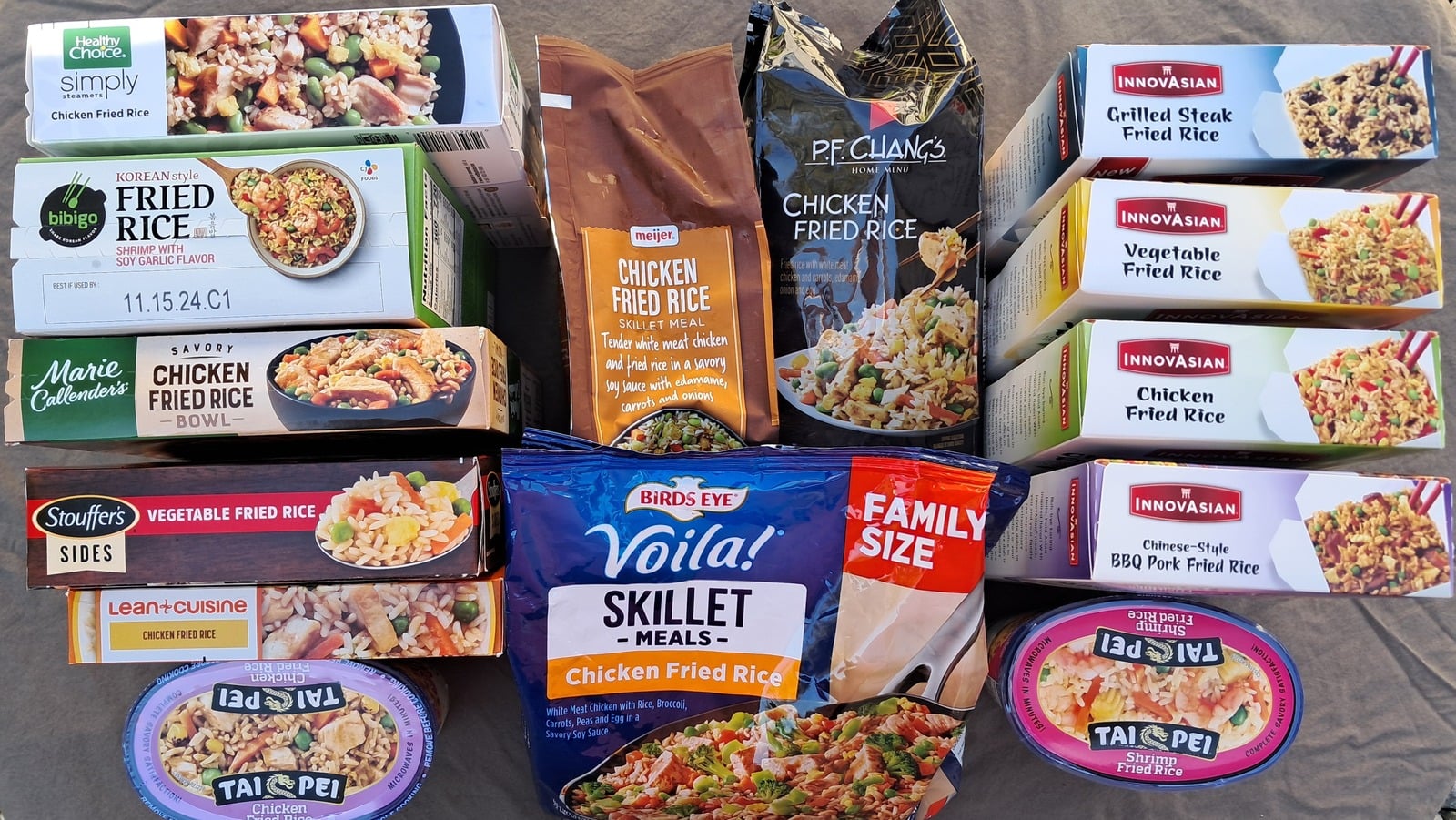 An assortment of 13 packaged frozen chicken fried rice meals from various brands, arranged on a table in multiple rows with colorful packaging and clear labeling.
