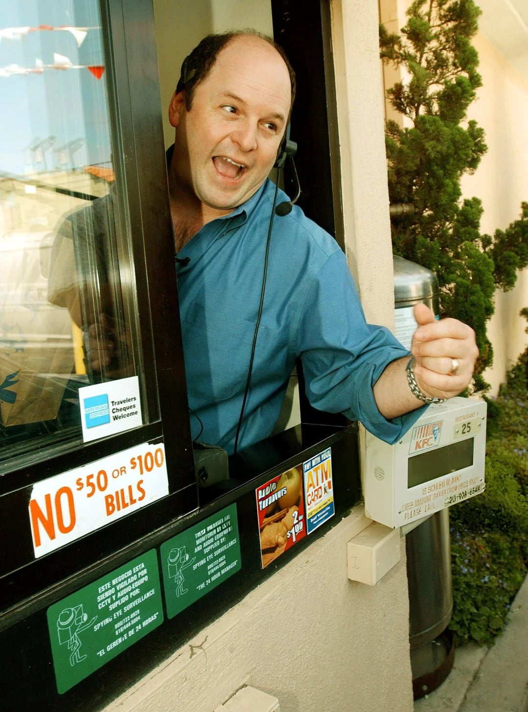 A cheerful man wearing a headset leans out of a fast-food drive-thru window, gesturing with his hand. Vintage Fast-Food Photos vibes are evident, with signs like "NO $50 OR $100 BILLS" and KFC branding visible near the window.