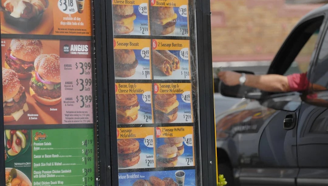 A drive-thru menu board displaying breakfast sandwiches and meal prices, with a person in a vehicle reaching out to the payment window—capturing the charm of vintage fast-food photos.