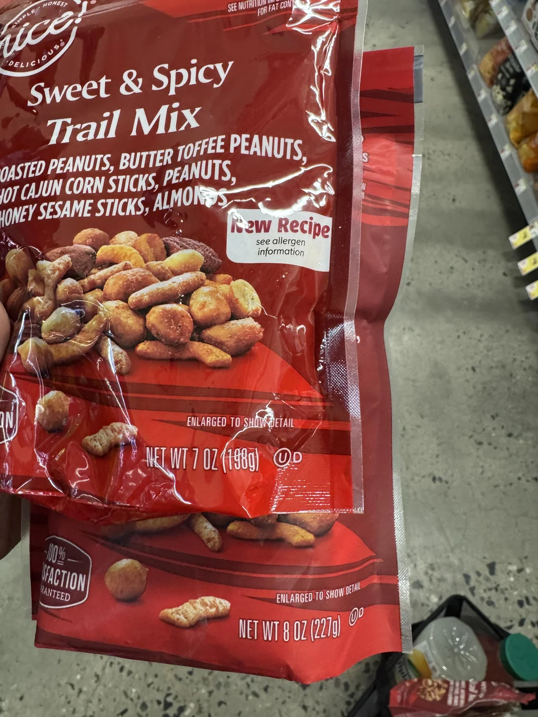 A hand holds a bag of Sweet & Spicy Trail Mix in a grocery store aisle. The bag displays images of the mix and shows the net weight as 7 oz (198g), while the bag below shows 8 oz (227g).