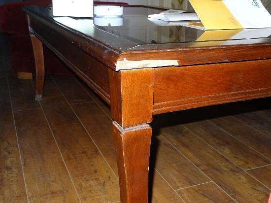 A wooden table with a glass top and visible damage to one corner, showing chipped and cracked wood. Papers and objects are placed on the tabletop. The floor is wooden, reflecting a style seen in emerging Hotel Brands 2026 interiors.