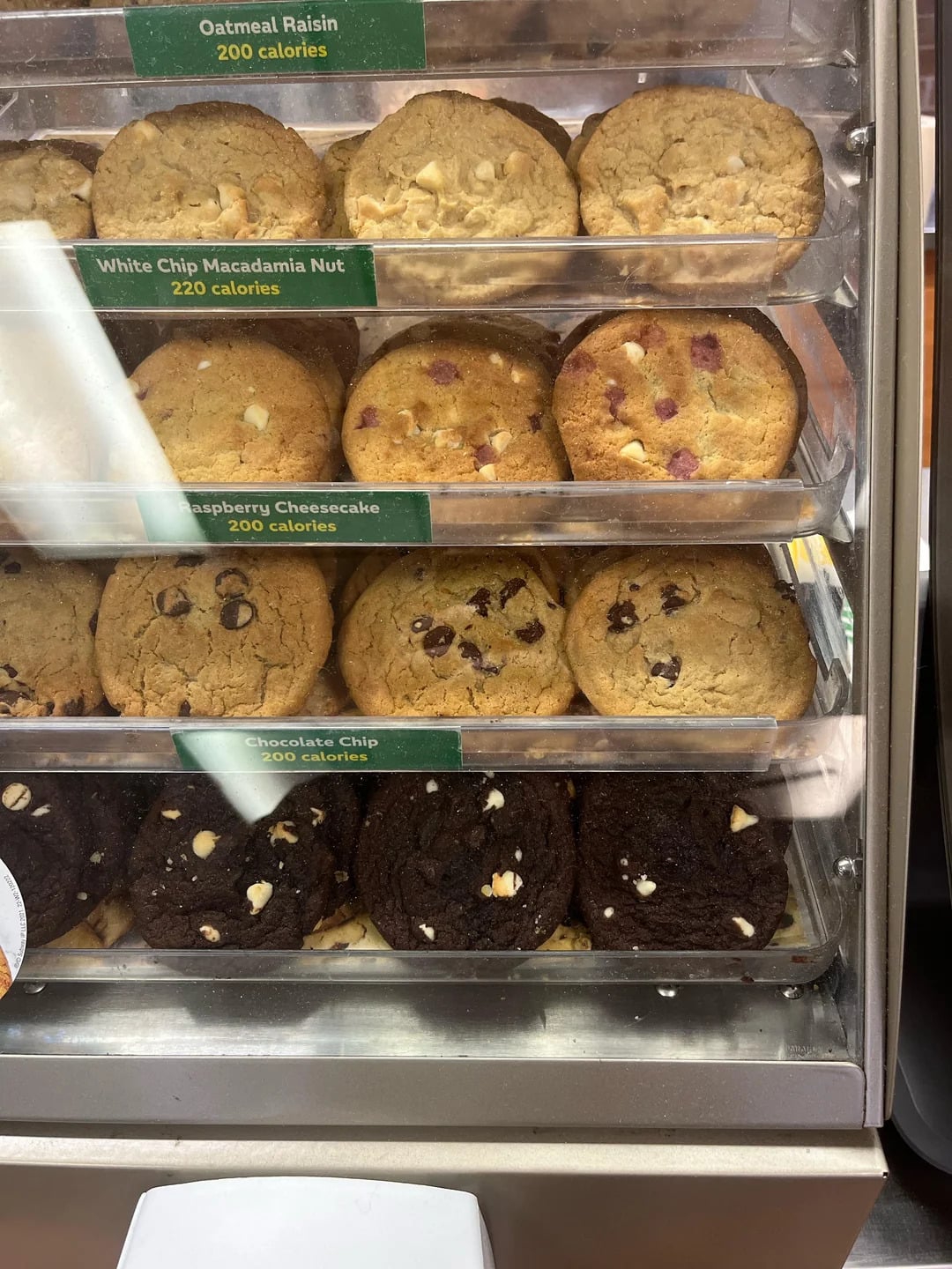A display case with four rows of cookies labeled as oatmeal raisin, white chip macadamia nut, raspberry cheesecake, and chocolate chip, each with a calorie count. The cookies are arranged neatly on trays.