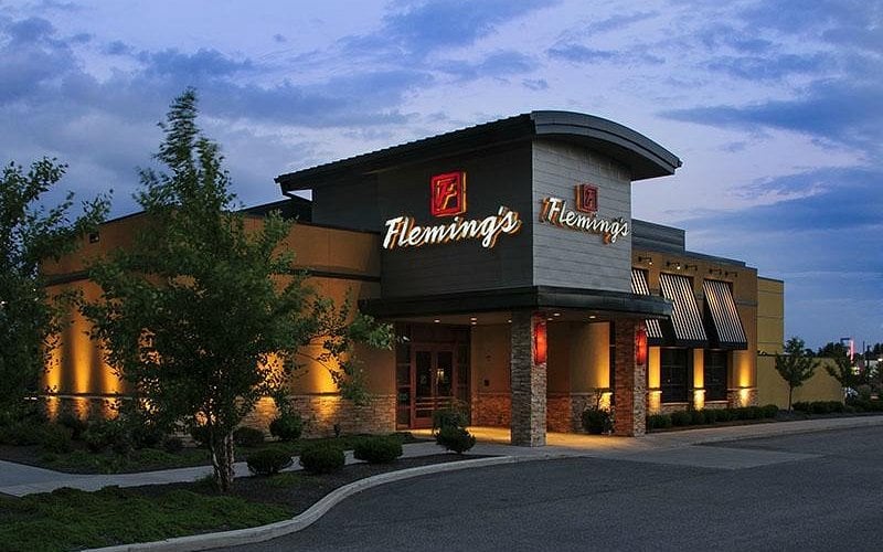 Exterior view of Fleming’s restaurant at dusk, with illuminated signs and warm lighting highlighting the modern building, stone accents, landscaped bushes, and a partly cloudy blue sky above.