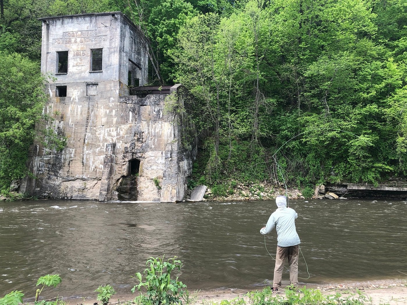 A person fly-fishing in a river stands across from an old, weathered concrete structure with faded writing, surrounded by dense, green trees—an authentic scene you might find in one of many Unique American Towns.