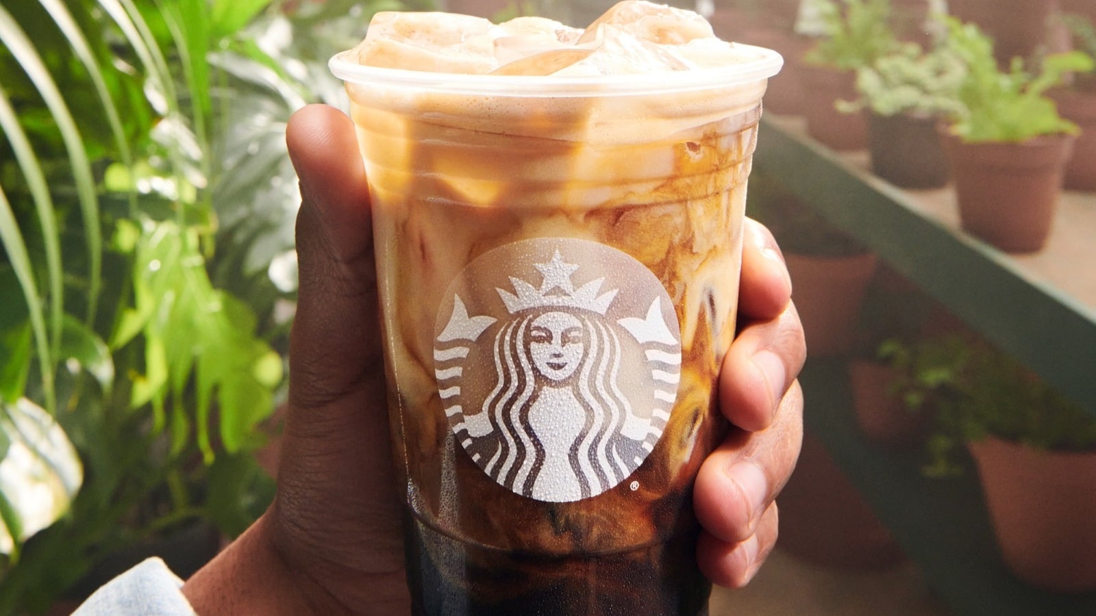 A hand holding a clear plastic Starbucks cup filled with iced coffee, showing swirls of milk and coffee, with a background of green plants and potted herbs.