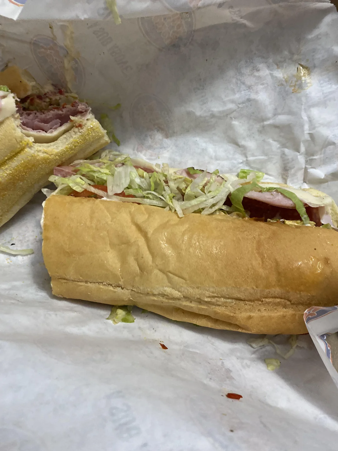 A sub sandwich with deli meat, shredded lettuce, and tomato on a hoagie roll is partially wrapped in white paper, with one half cut open and some lettuce scattered on the paper.