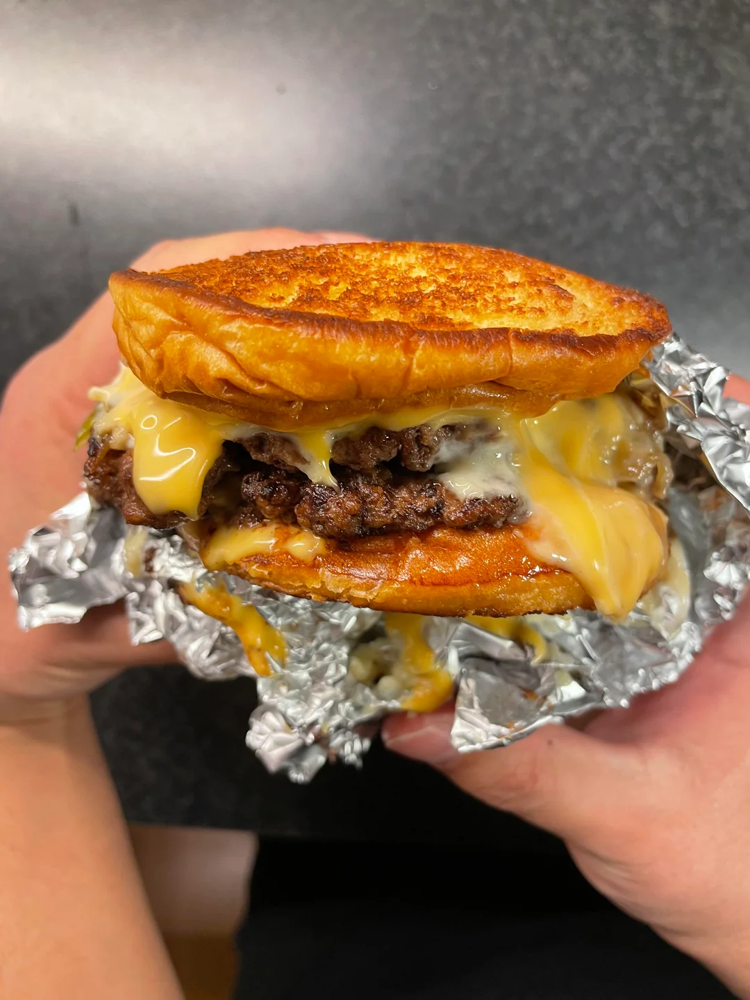 A person holds a large, cheesy double cheeseburger with a toasted bun, wrapped partially in aluminum foil. Melted cheese drips over the juicy beef patties, with a dark countertop in the background.