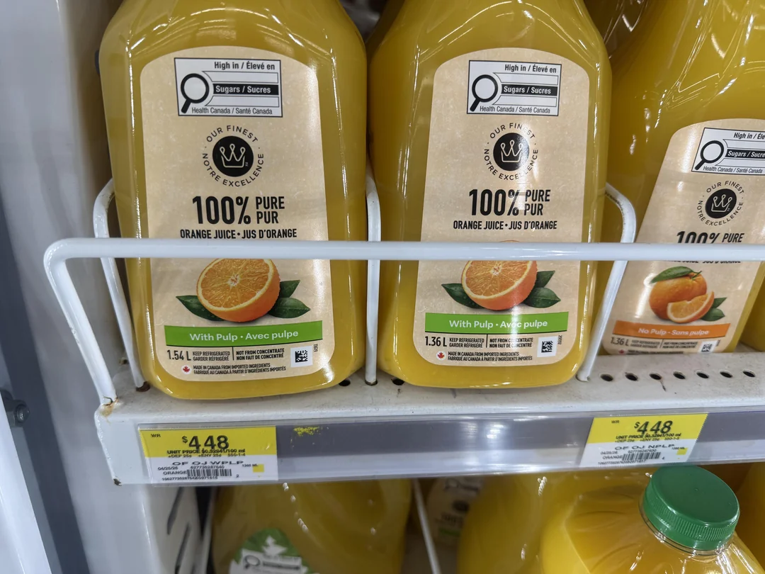 Bottles of 100% pure orange juice with pulp are displayed on a store shelf. The yellow bottles are labeled 1.54 L and 1.36 L, with price tags reading $4.48 and $4.28.