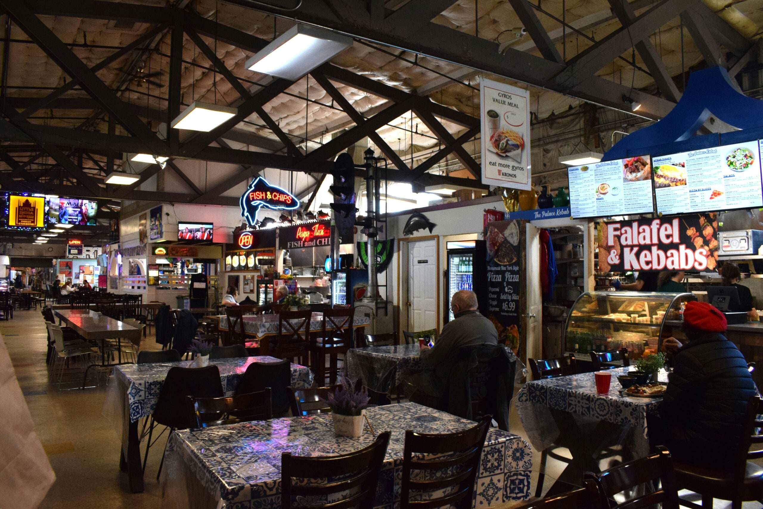 A food court with tables and chairs, various food stalls including "Falafel & Kebabs" and "Fish & Chips," and people sitting and eating. Bright signs and menus are visible under exposed ceiling beams.