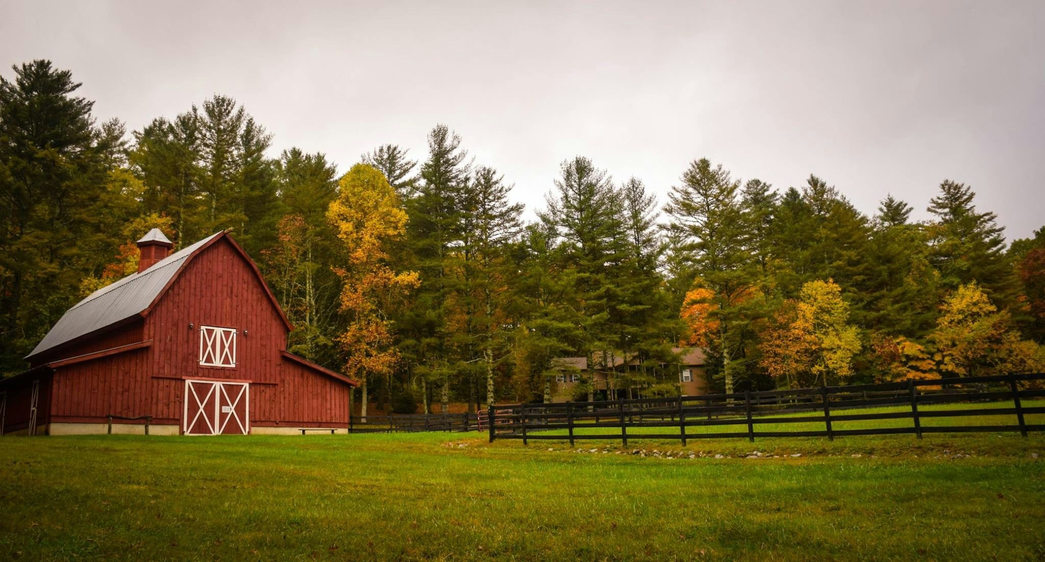 A red barn with white trim sits on green grass, bordered by a black wooden fence. Trees with green and autumn-colored leaves stand behind the barn under a cloudy sky.