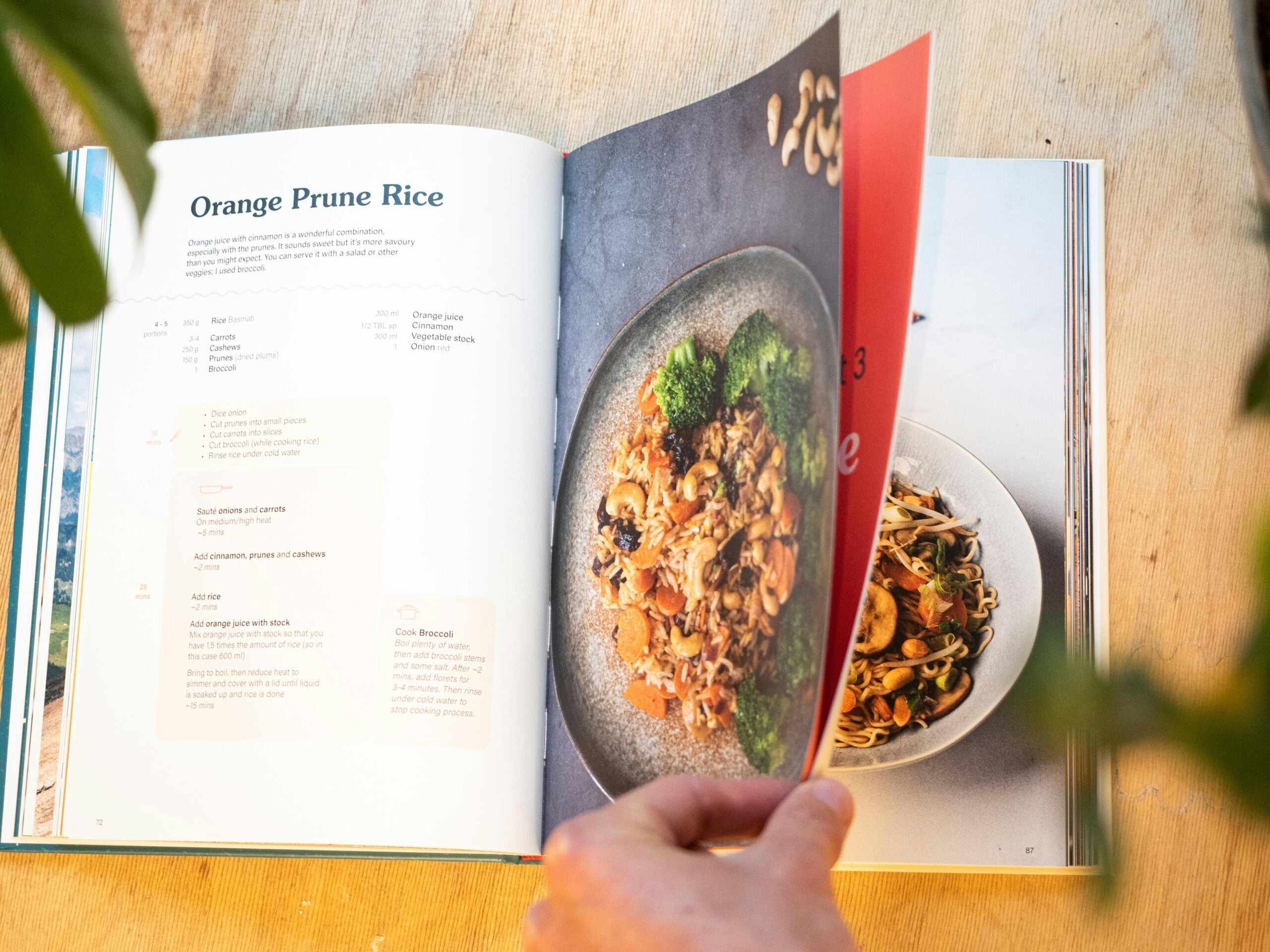A hand flips a page of a cookbook, revealing a recipe for Orange Prune Rice on the left and a photo of a plated rice dish with vegetables on the right. The book rests on a wooden surface.