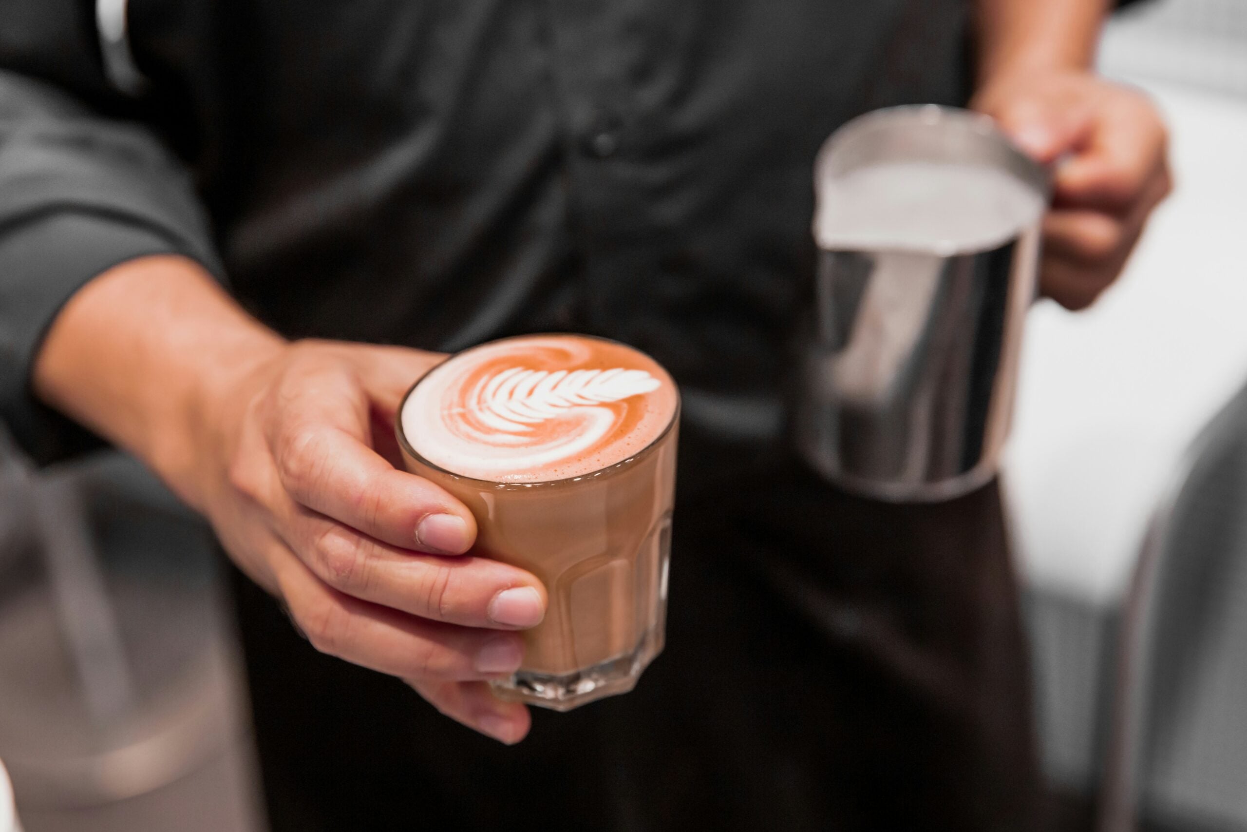 A person holding a glass of coffee with latte art in one hand and a metal milk frothing pitcher in the other, wearing a dark shirt.